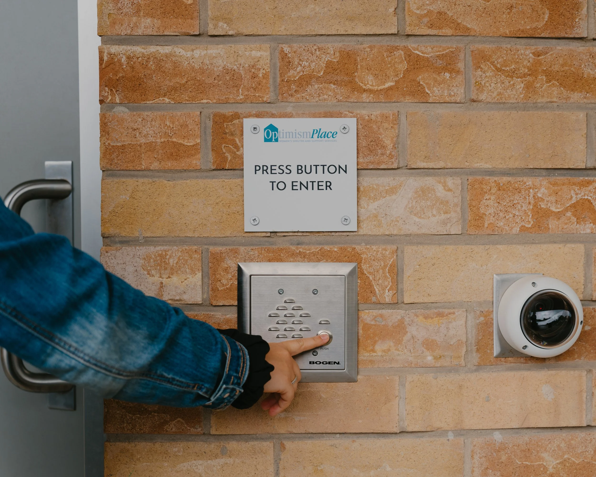 Person pressing a button on a security keypad next to a sign that says 'Press Button to Enter' at a building entrance.