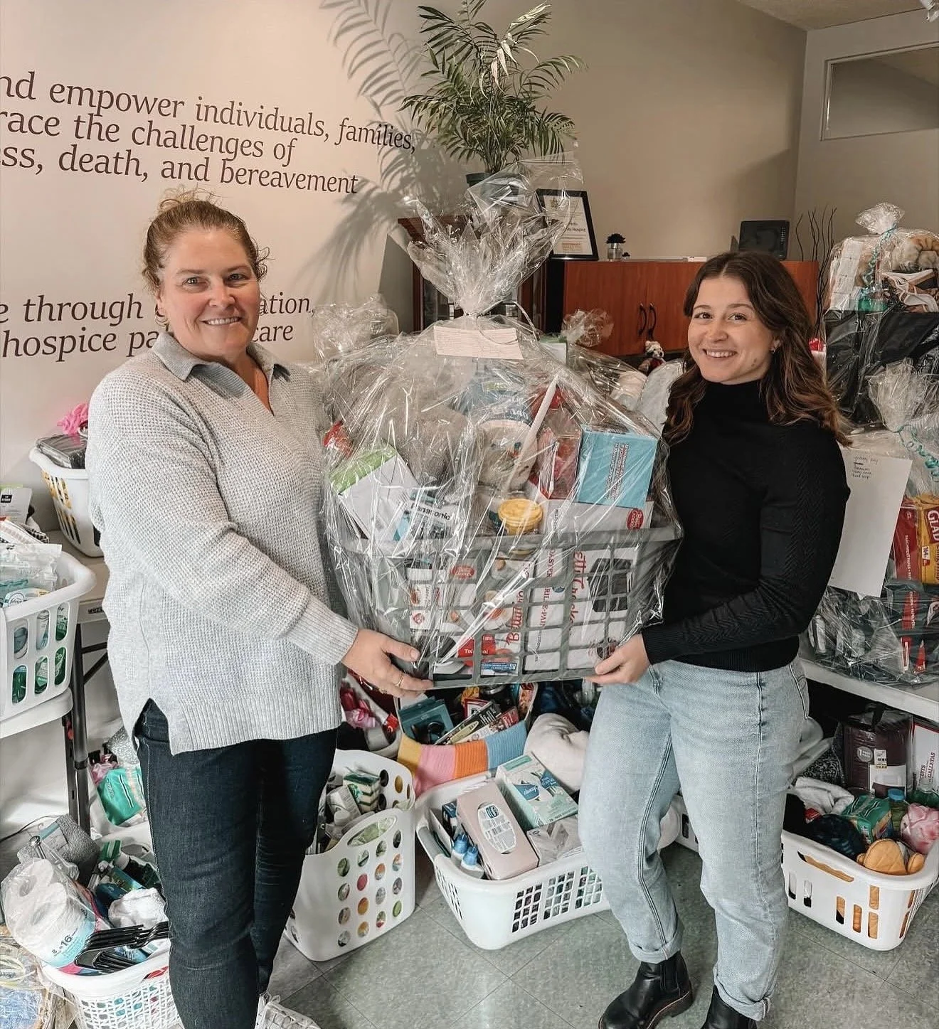 Two women smiling and holding a large wrapped gift basket filled with various items, in a room with baskets of supplies around them.