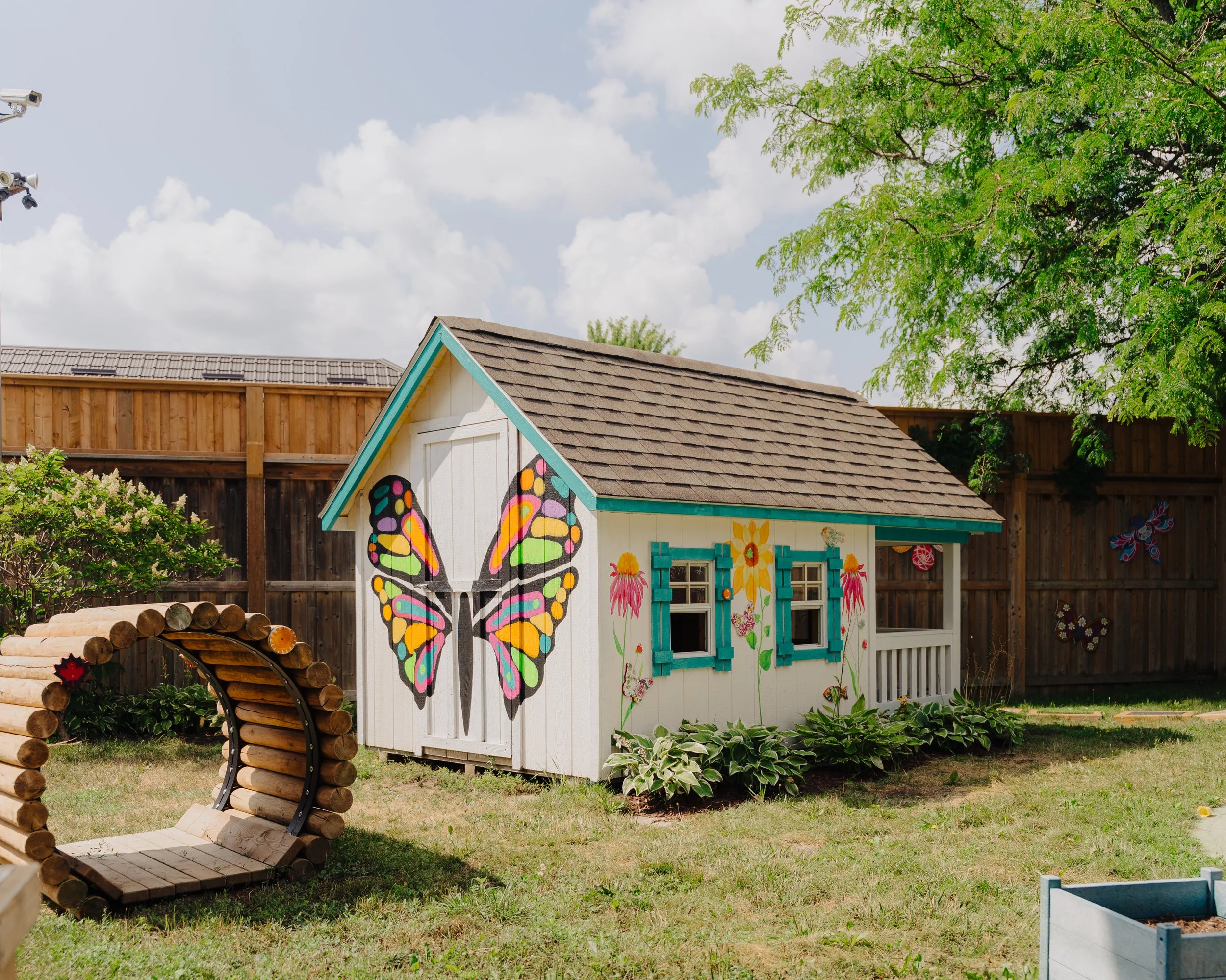 A small garden shed painted white with blue trim, decorated with colorful butterfly and flower murals, sitting on a grassy yard with a wooden fence and trees in the background.