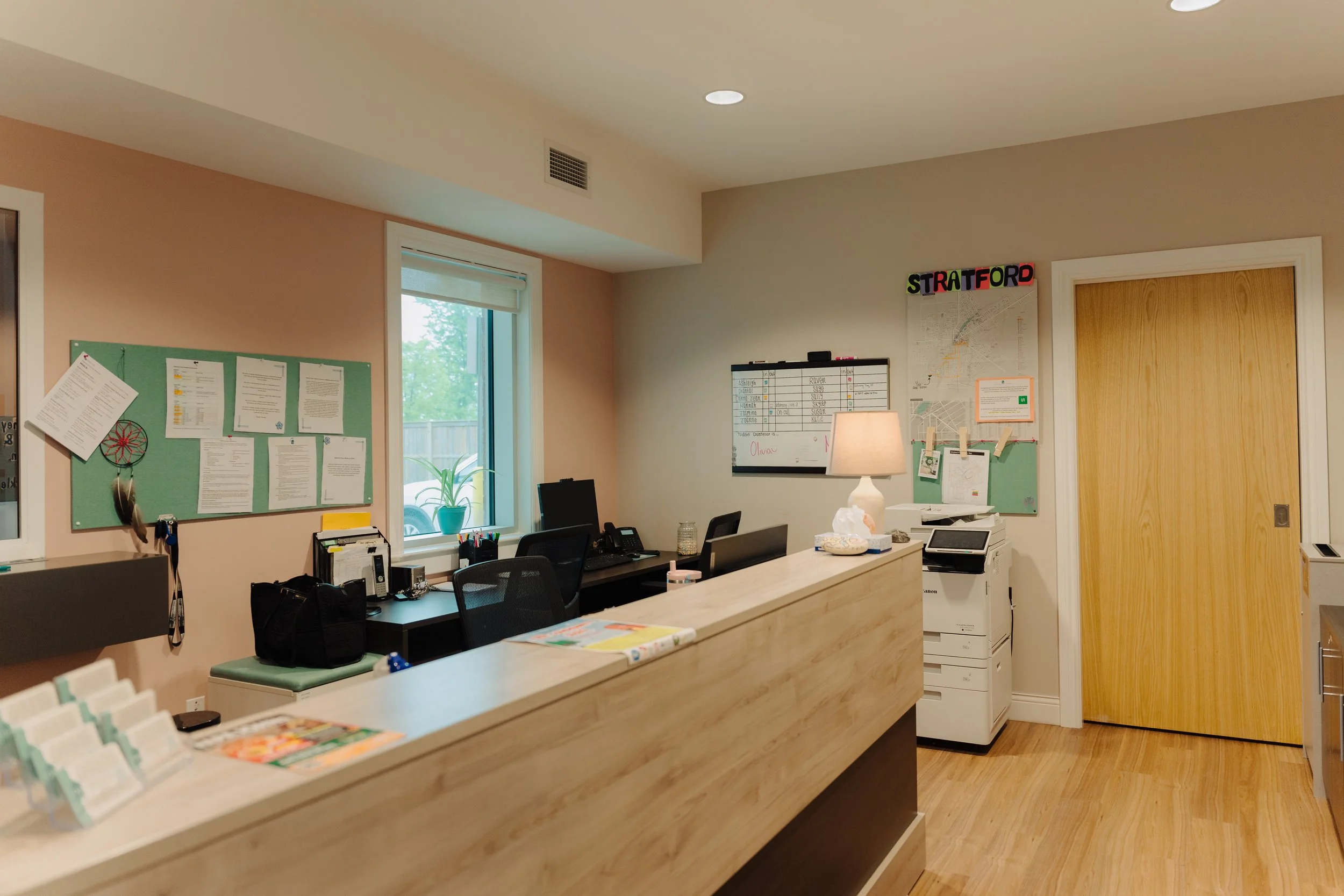 Interior view of an office with a long reception desk, a window with a plant outside, a bulletin board, a whiteboard, a bookshelf, computers, and a closed wooden door.