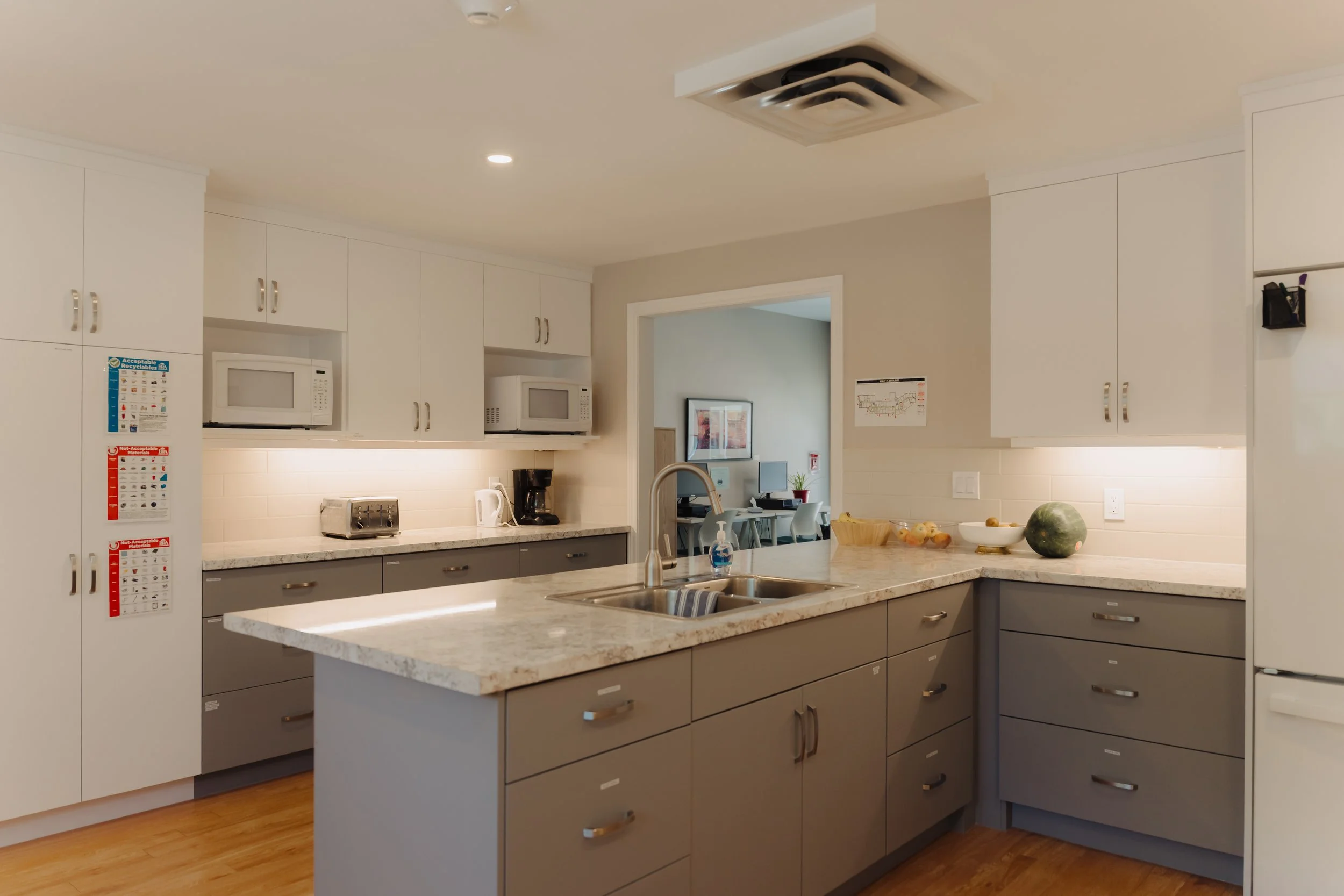 Modern kitchen with gray cabinets, white upper cabinets, granite countertops, and small appliances on the counter, with a view into a dining or living area with a desk and computer in the background.