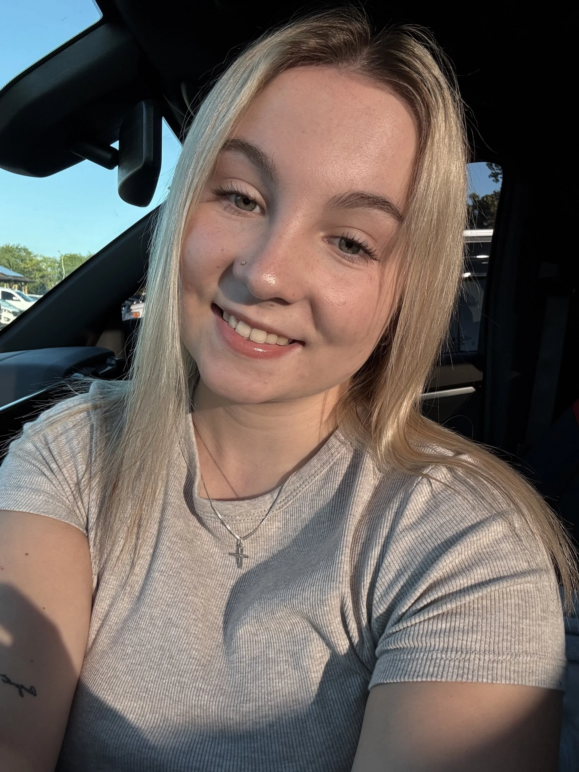 A young woman with blonde hair taking a selfie inside a car, smiling, wearing a beige shirt and a necklace with a cross pendant, with sunlight shining on her face.