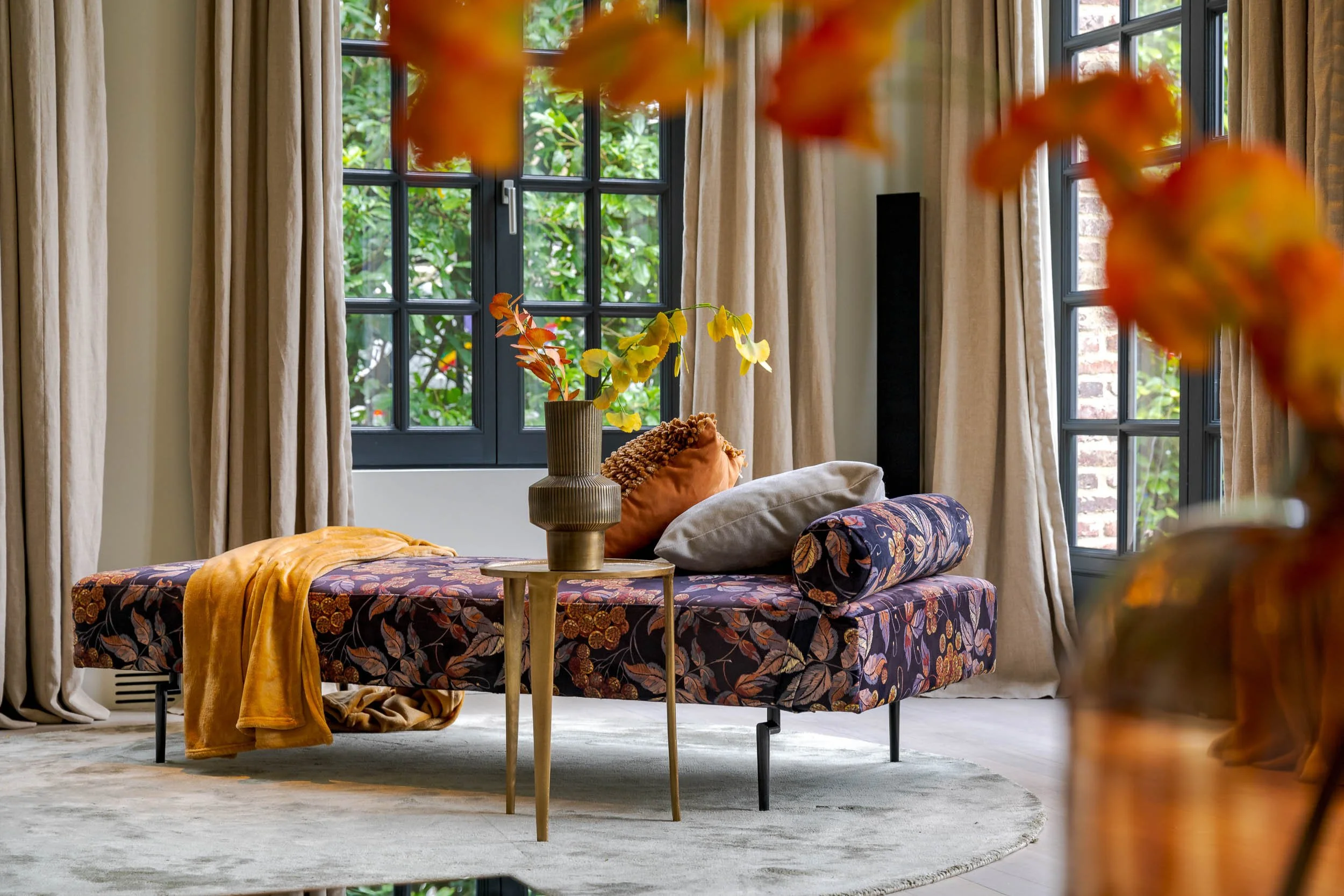 Interieur van een villa in Wassenaar woningfotografie van een woonkamer met grote ramen, beige gordijnen, een gestoffeerde bank met bloemendessin, een ronde tafel met een vaas met herfstbladeren, en een zachte grijze vloerbedekking.