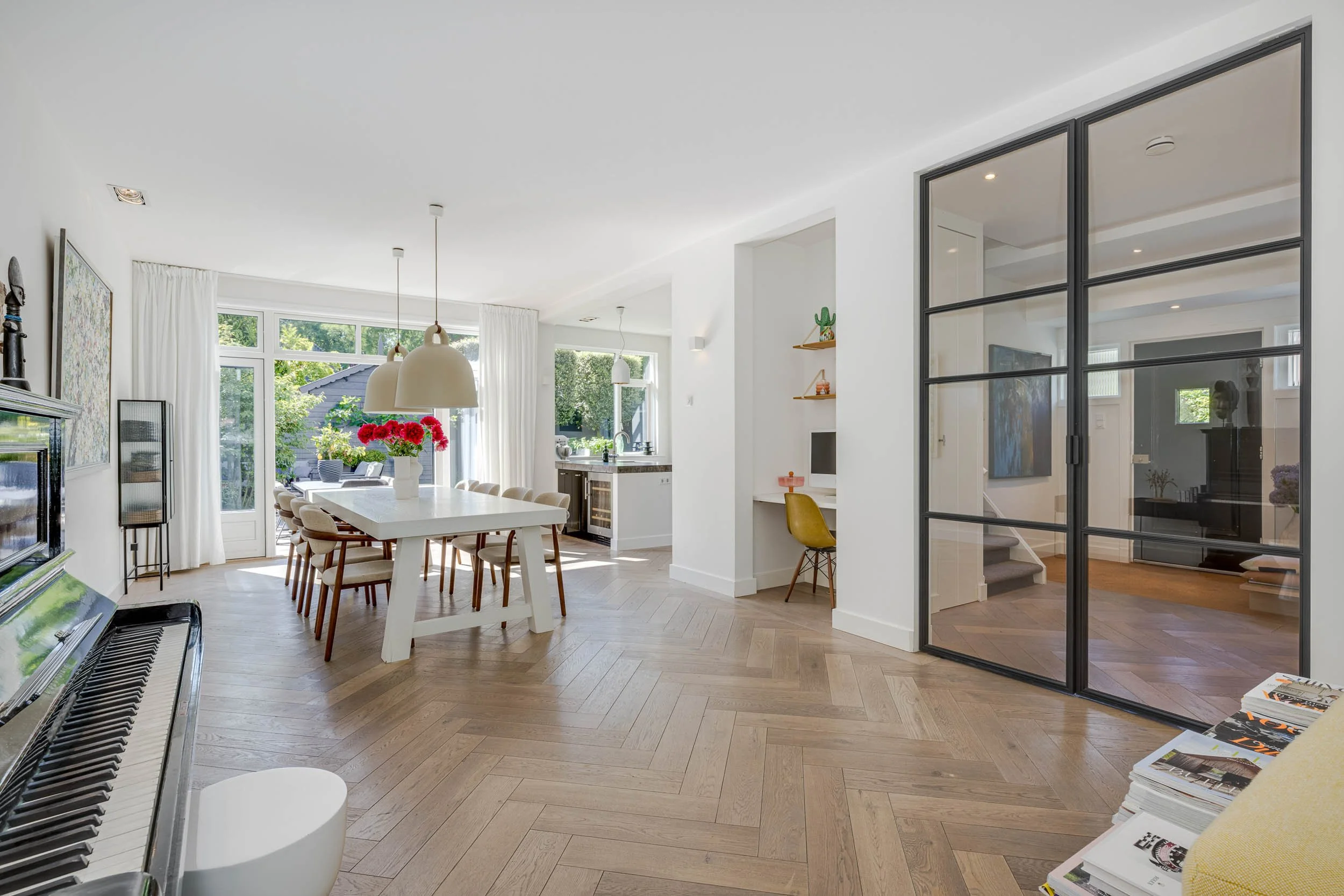 Interieur van een modern huis met een eettafel en stoelen, grote ramen met witte gordijnen, getegelde vloer, piano, en een grote glazen schuifdeur die naar een tuin leidt.
