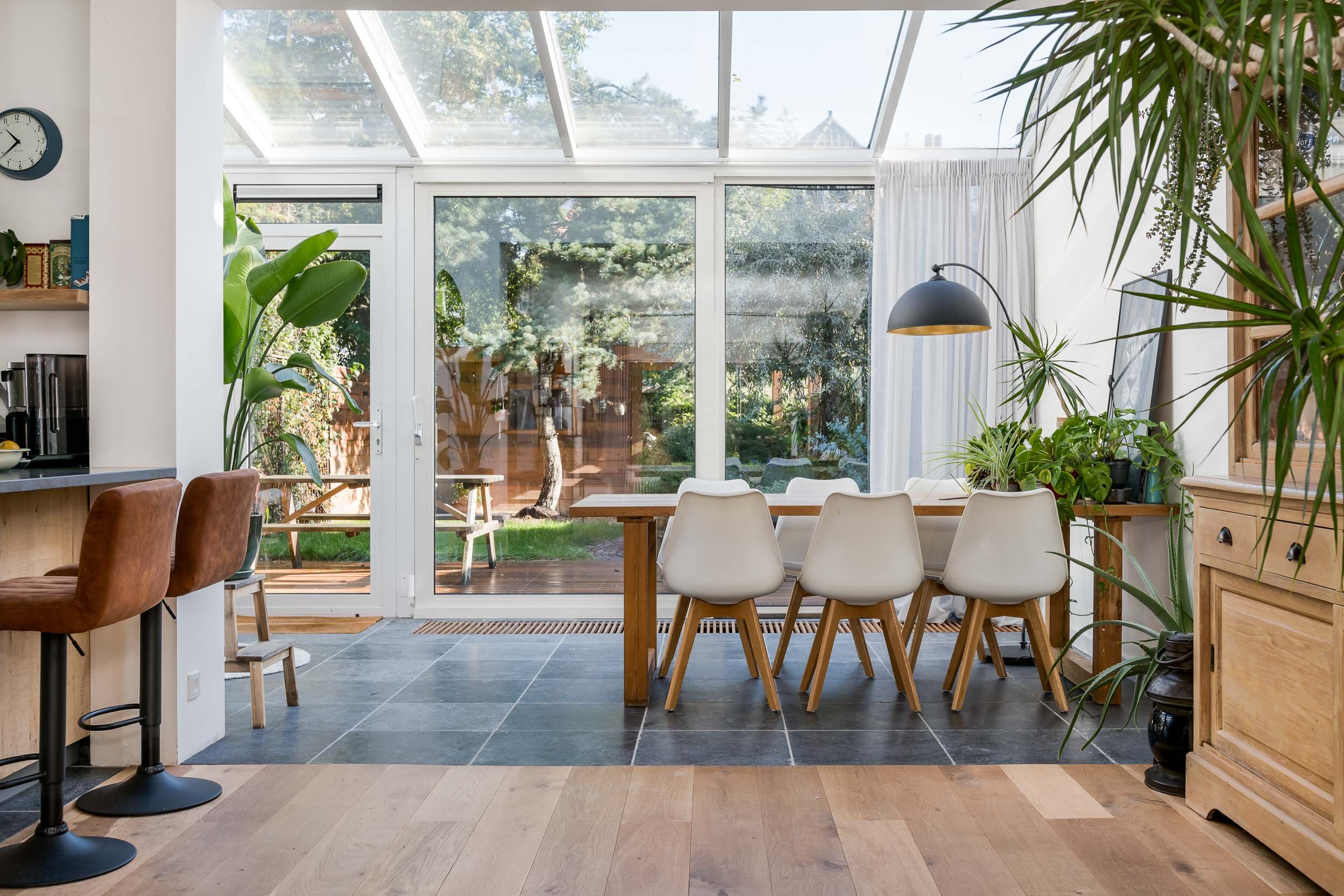 Een eetkamer met een grote tafel en witte stoelen, uitzicht op een tuin via grote glazen schuifdeuren, groene planten binnen, een zwarte hanglamp boven de tafel.