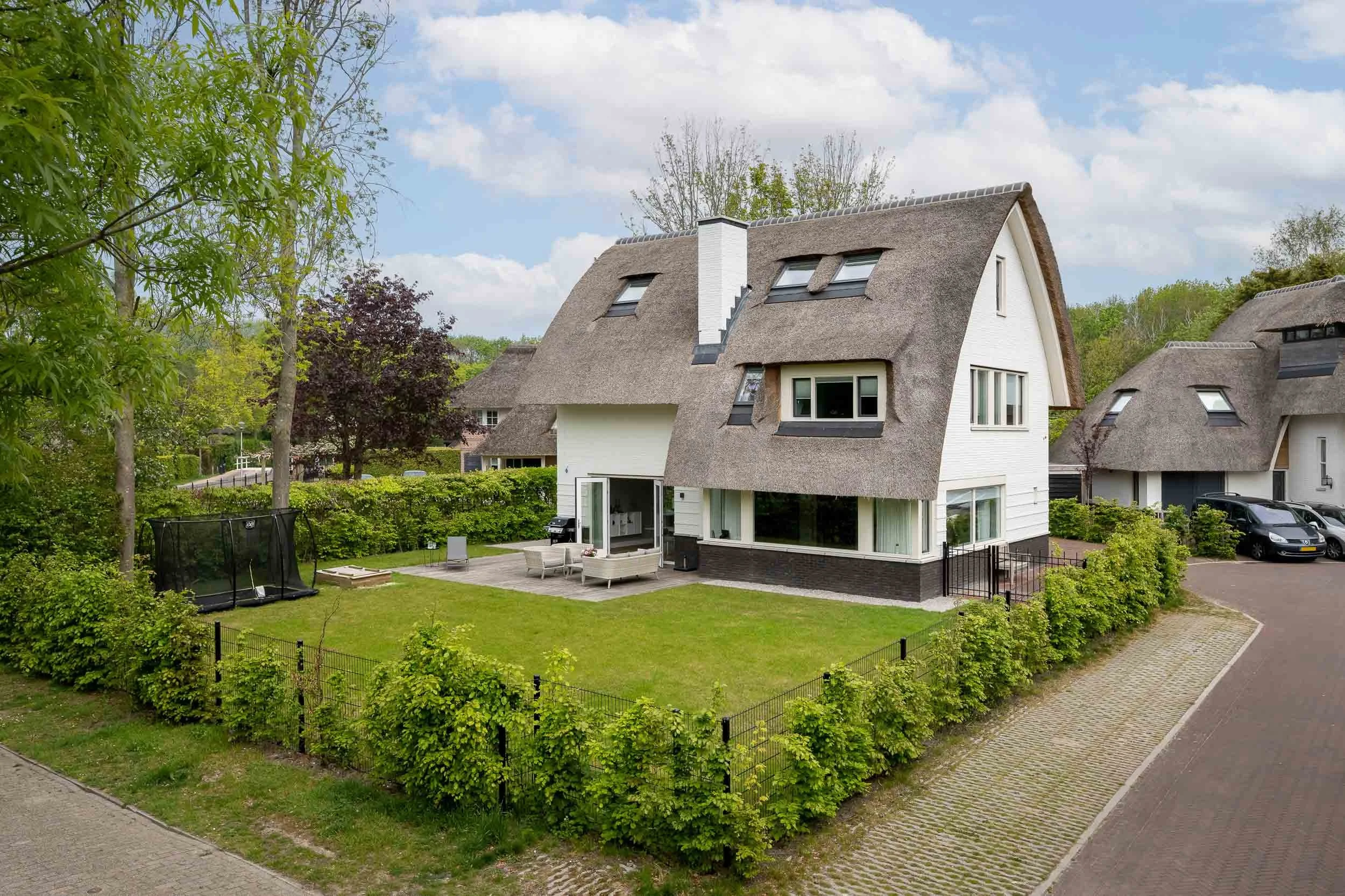 Een modern huis met een rieten dak en grote ramen, omgeven door een groene tuin met bomen en een klein terras met zitplaatsen.