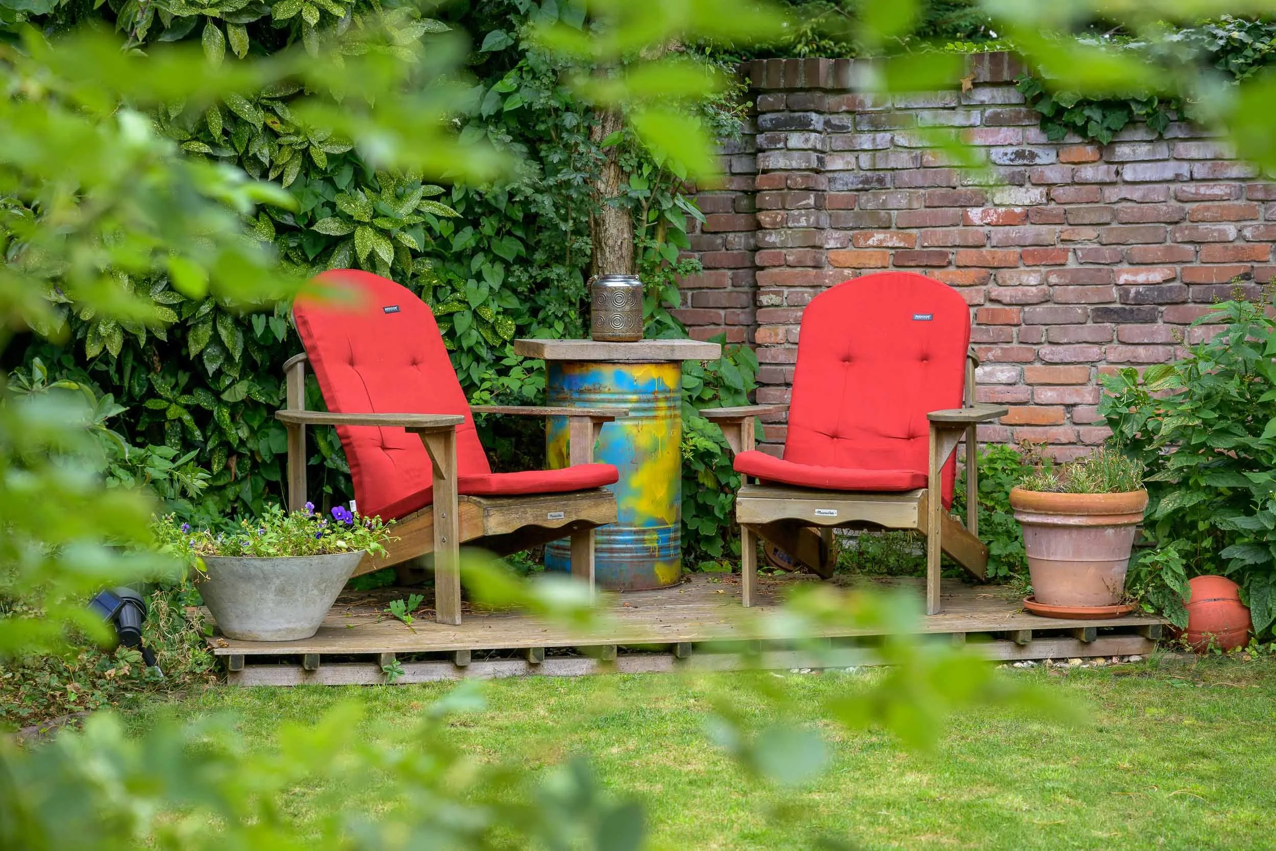 Twee rode tuinligstoelen op een houten terras met planten en struiken, naast een bak met bloemen en een oude metalen minicontainer op een magere tafel, een stenen muur op de achtergrond.