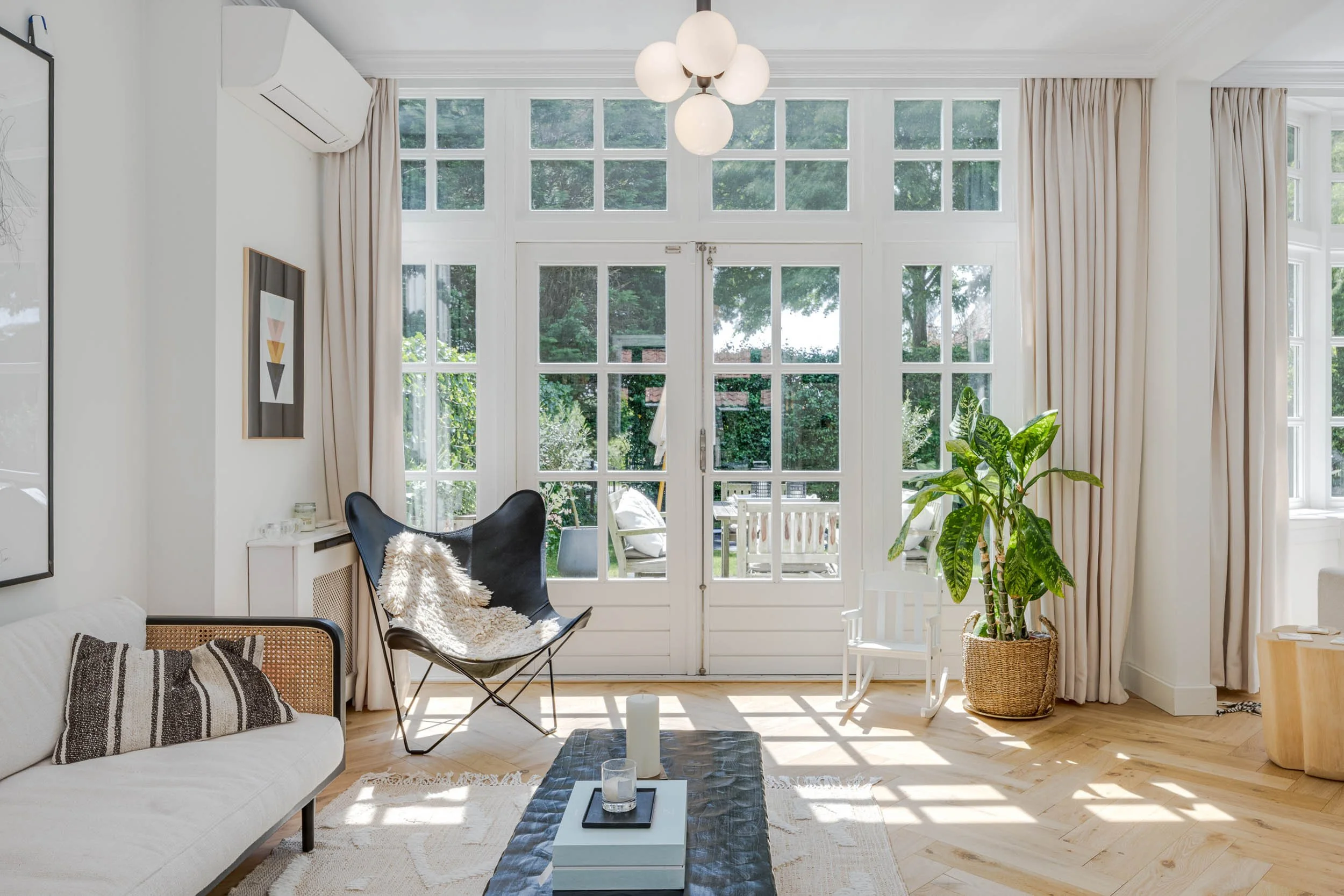 Een woonkamer met grote ramen en beige gordijnen, een plant in een mand, een zwarte en witte stoel met een wollen deken, een bank met kussen, en een tafel met kaarsen en glazen.