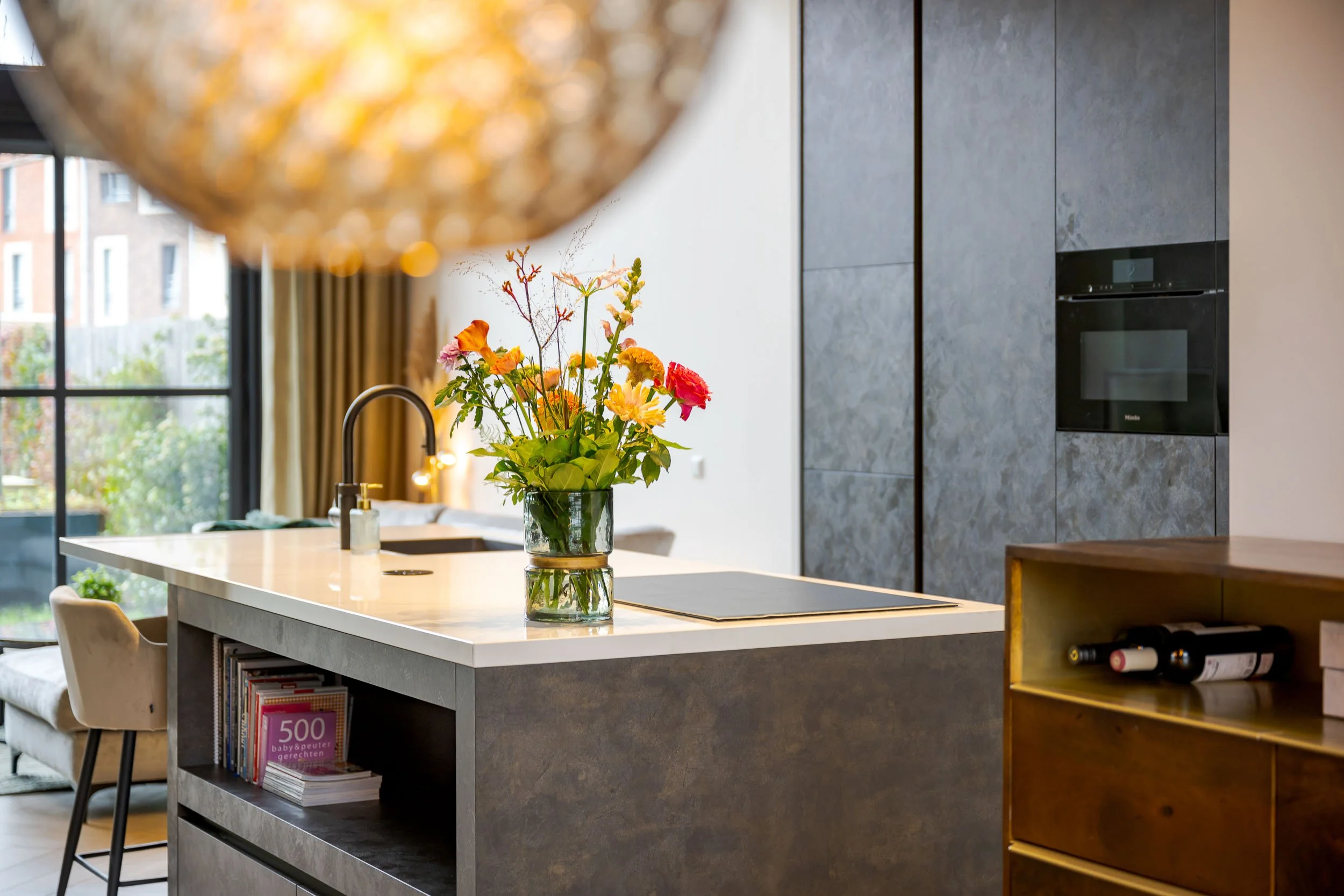 Binnenkant van een moderne keuken met een lichtroze keukenblad en een bloempot met kleurrijke bloemen op het aanrecht, grote ramen met uitzicht op gebouwen, en een ingebouwde oven in de muur.
