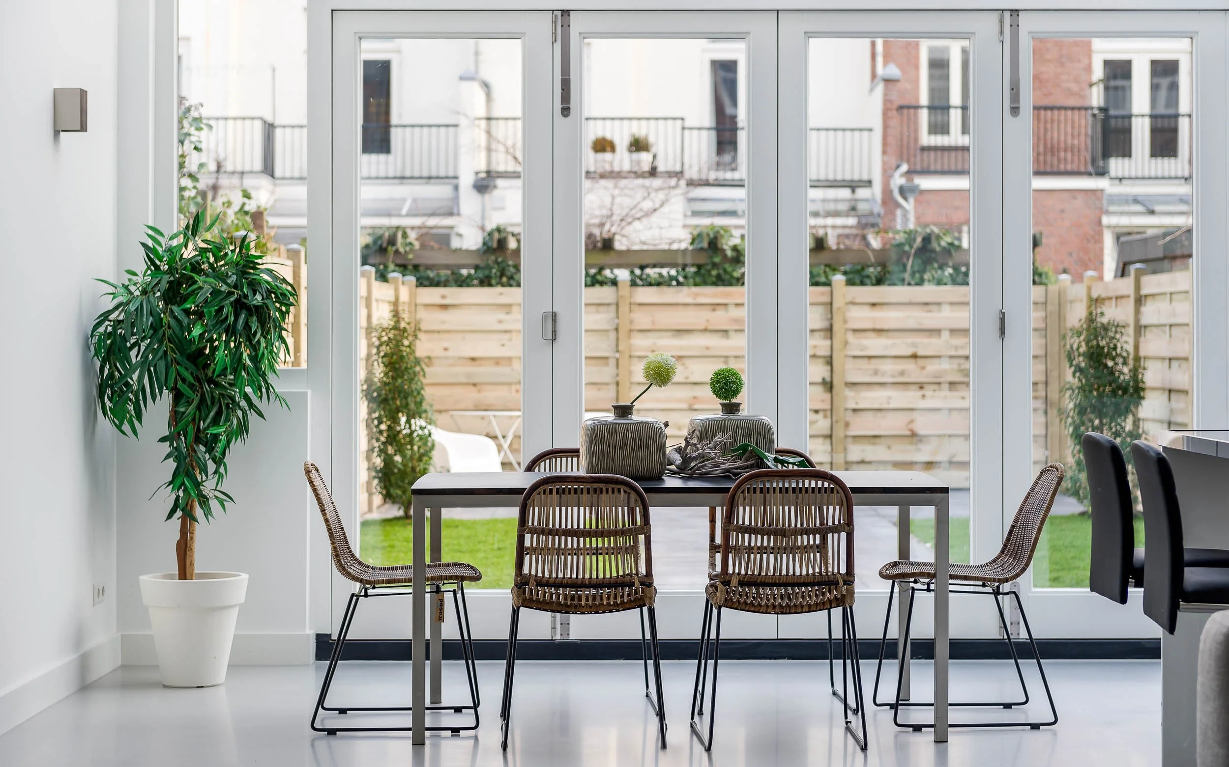 Binnenzijde van een woning met een eettafel en stoelen, uitzicht op een tuin met houten schutting en flats in de achtergrond.