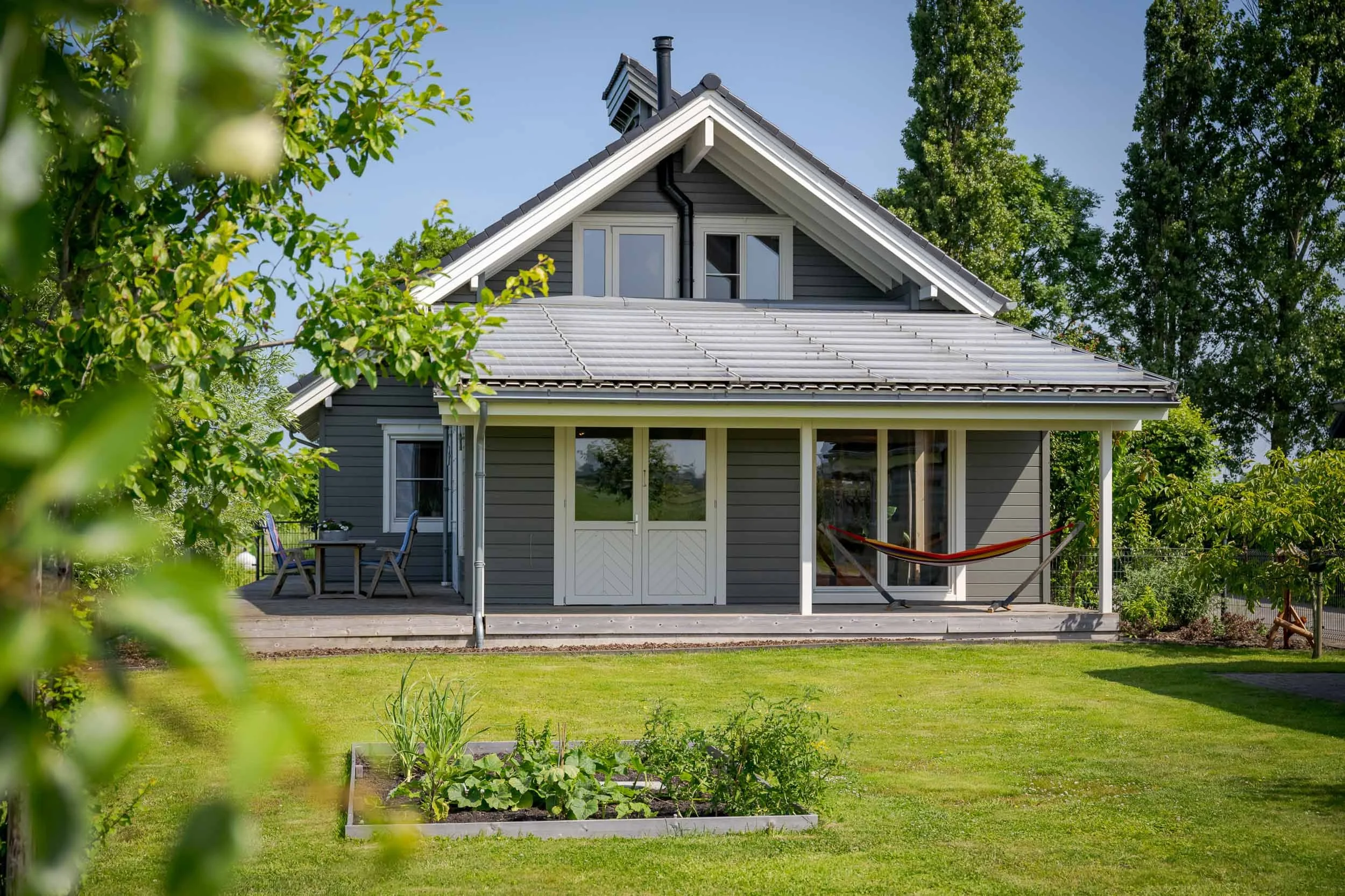 Een huis met een veranda, omringd door een groene tuin met een moestuin en bomen, onder een heldere blauwe hemel.