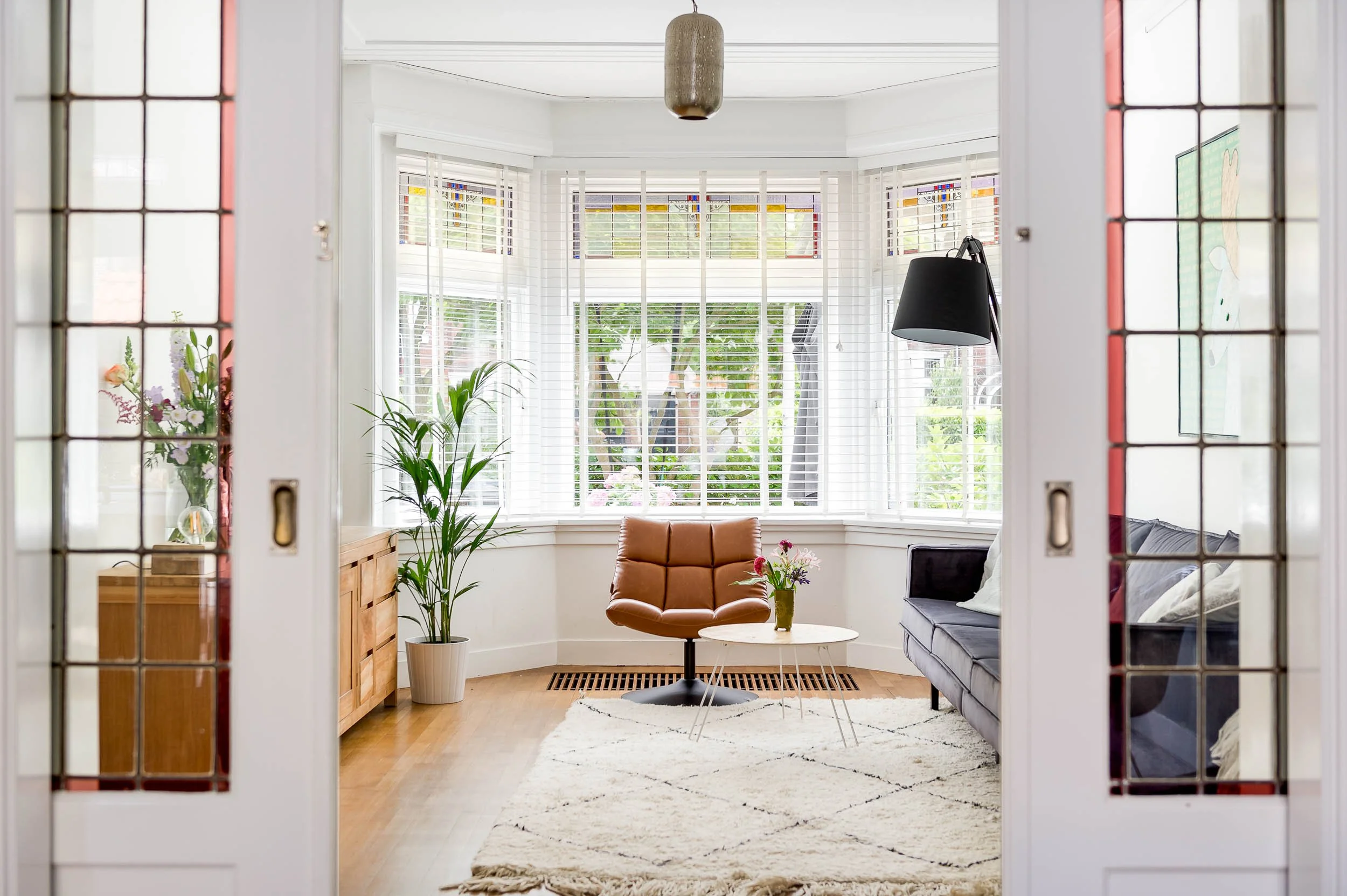 Een woonkamer met grote ramen en jaloezieën, een bruine leren stoel, een salontafel met vaas bloemen, een bank en planten.
