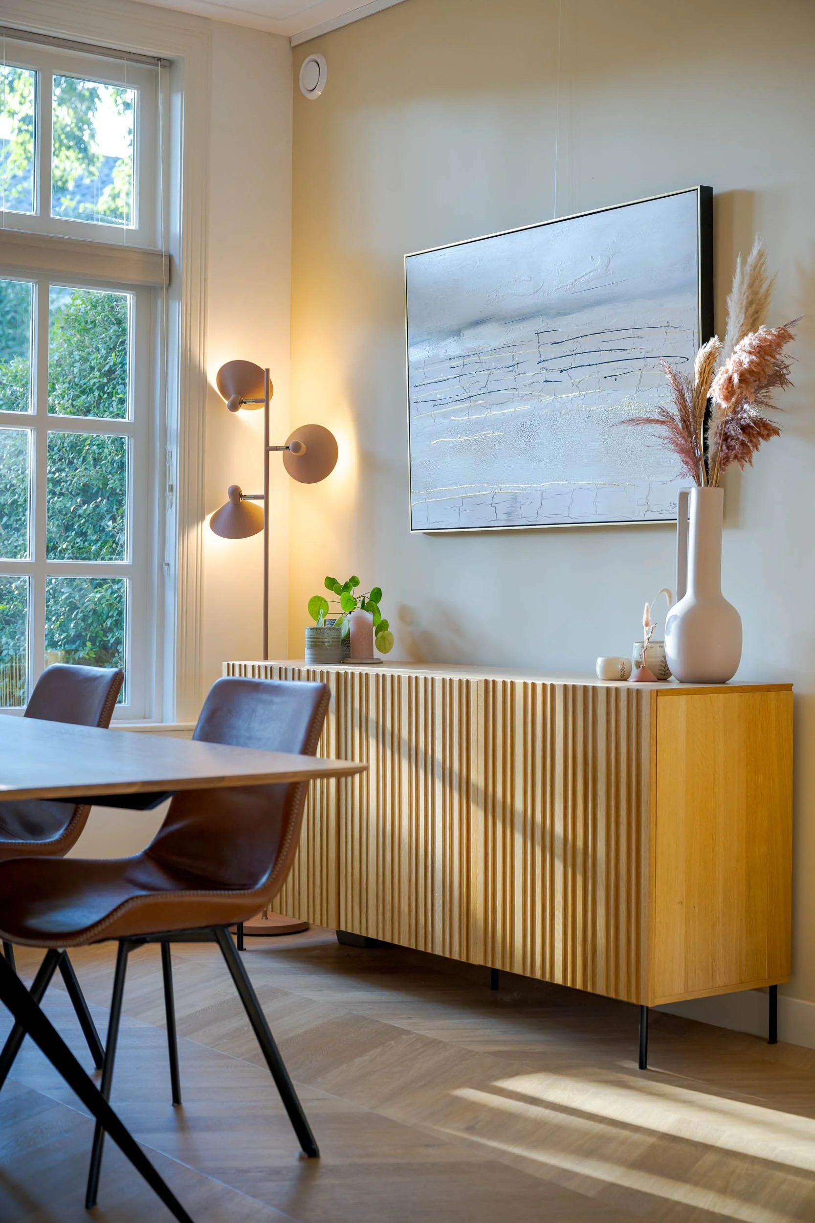 Interieur van een woonkamer met een grote witte vaas met gedroogde bloemen, een houten dressoir met planten en decoratie, een moderne lamp en een groot abstract schilderij aan de muur, naast een raam met uitzicht op groen.