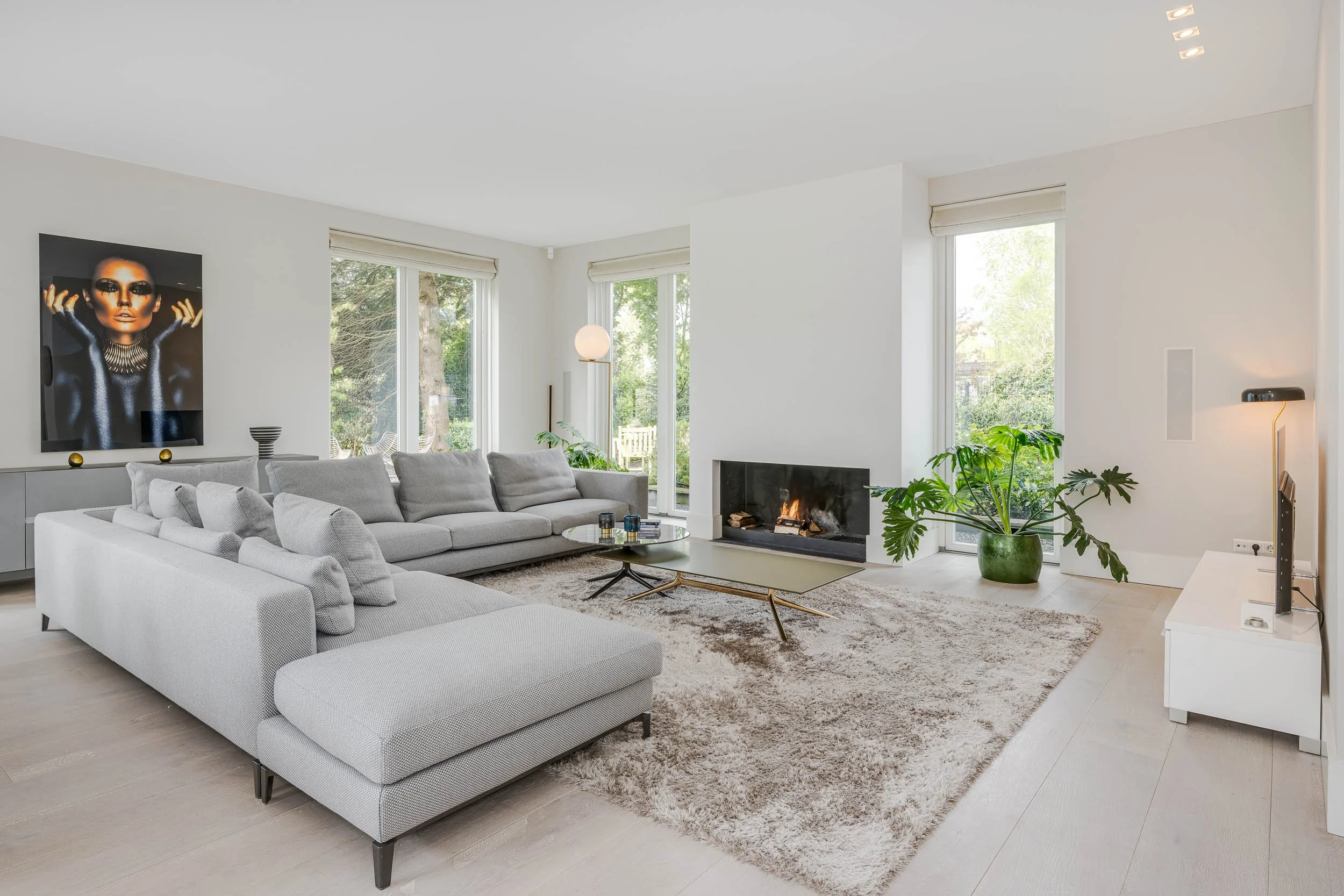 Een modern woonkamer met grote ramen, een witte muur, een lichtgrijze hoekbank, een zwarte salontafel, een donkerstenen haard, een grote groene plant, een schilderij van een vrouw met donkere make-up, een ronde witte lamp, en een beige vloerbedekking