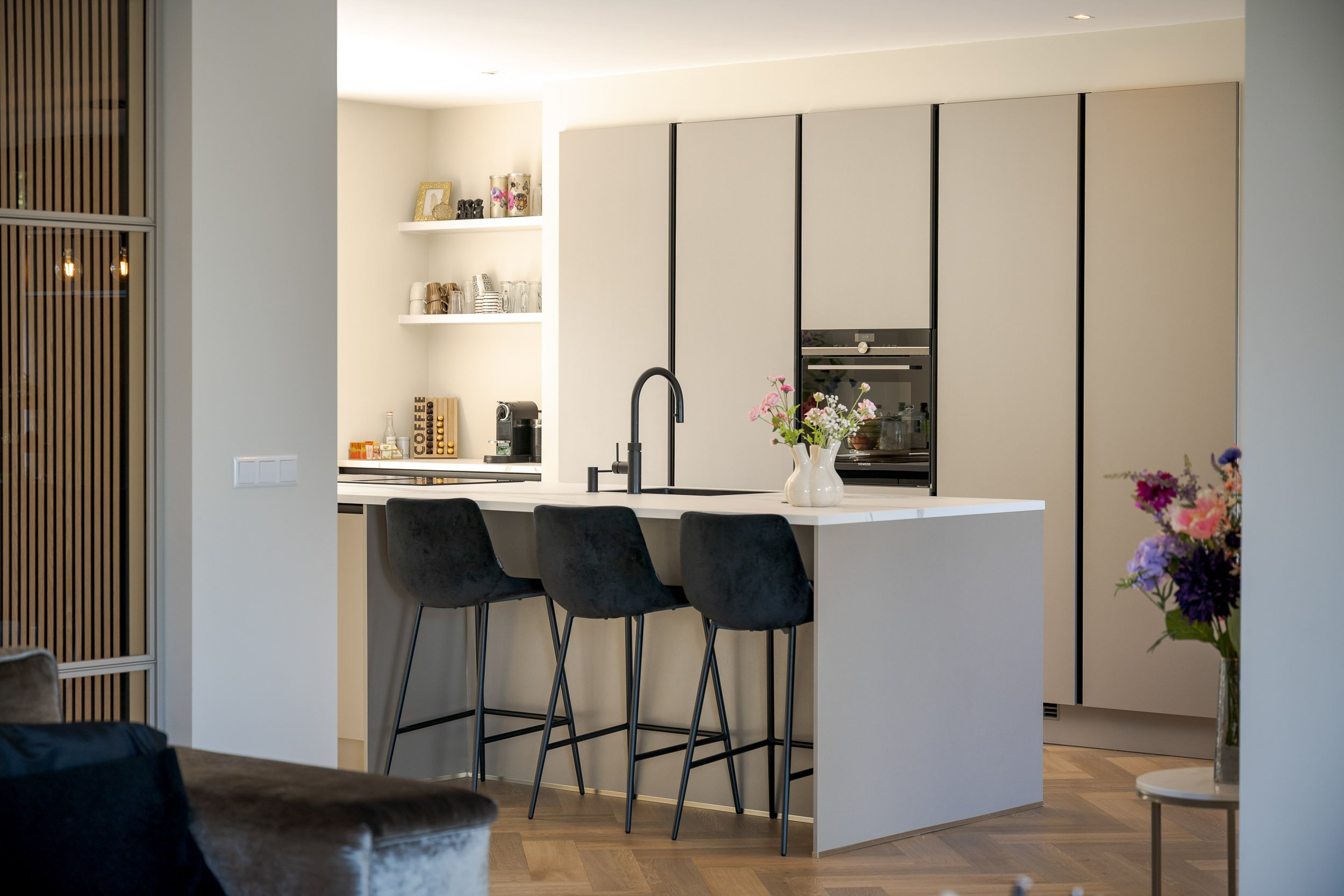 Een moderne keuken met een wit aanrechtblad, zwarte kranen, een bloemstuk in een witte vaas, en ingebouwde keukentoestellen, in een open woonruimte.