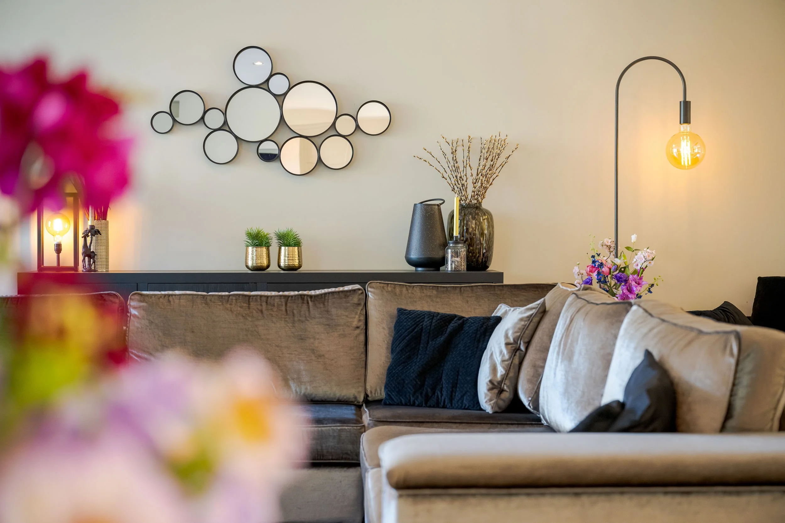 Een interieur met een beige bank, decoratieve kussens, een zwarte console met planten en vazen, een wandspiegel met meerdere cirkels, een vloerlamp met een gloeilamp, en een bloemstuk.