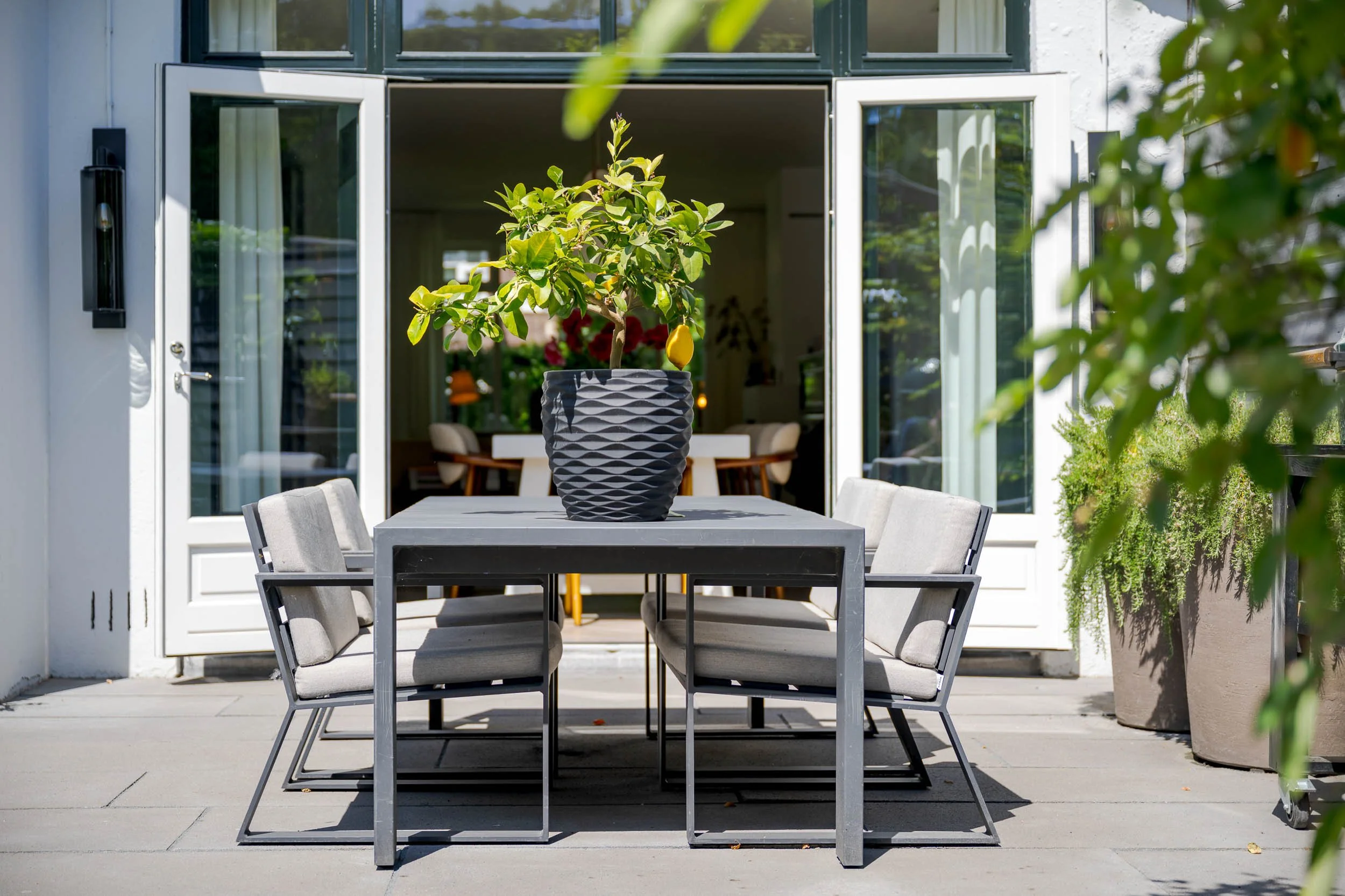 Buiten eethoek met een tafel en zes stoelen, met een pot met een groene plant en een citroen op de tafel, voor een huis met grote ramen en een open deur, in de tuin.