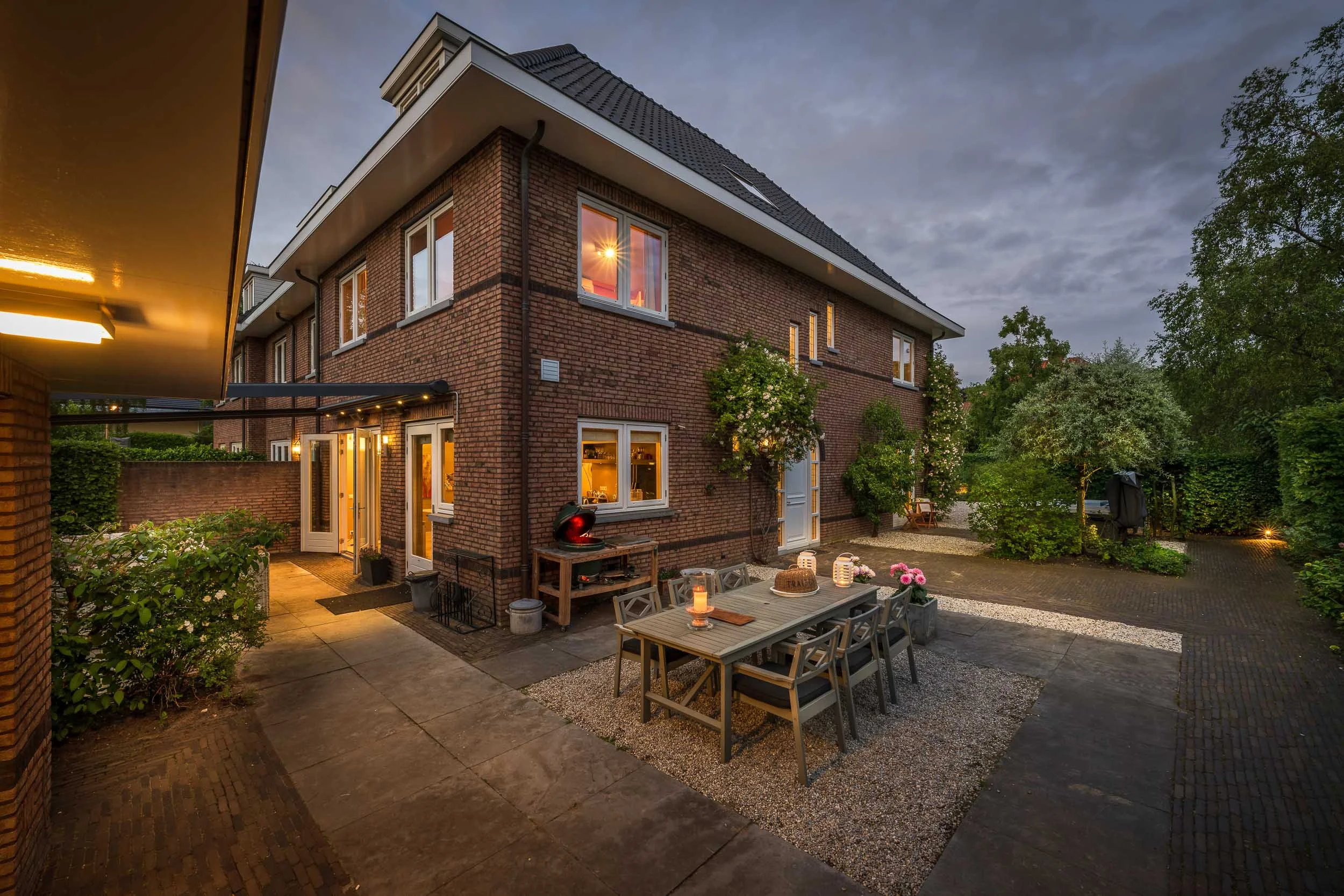 Achtertuin van een bricken woning met verlichte binnenkant en buitenverlichting, terras met tuinmeubilair, bloemen, en omringende bomen, 's avonds.