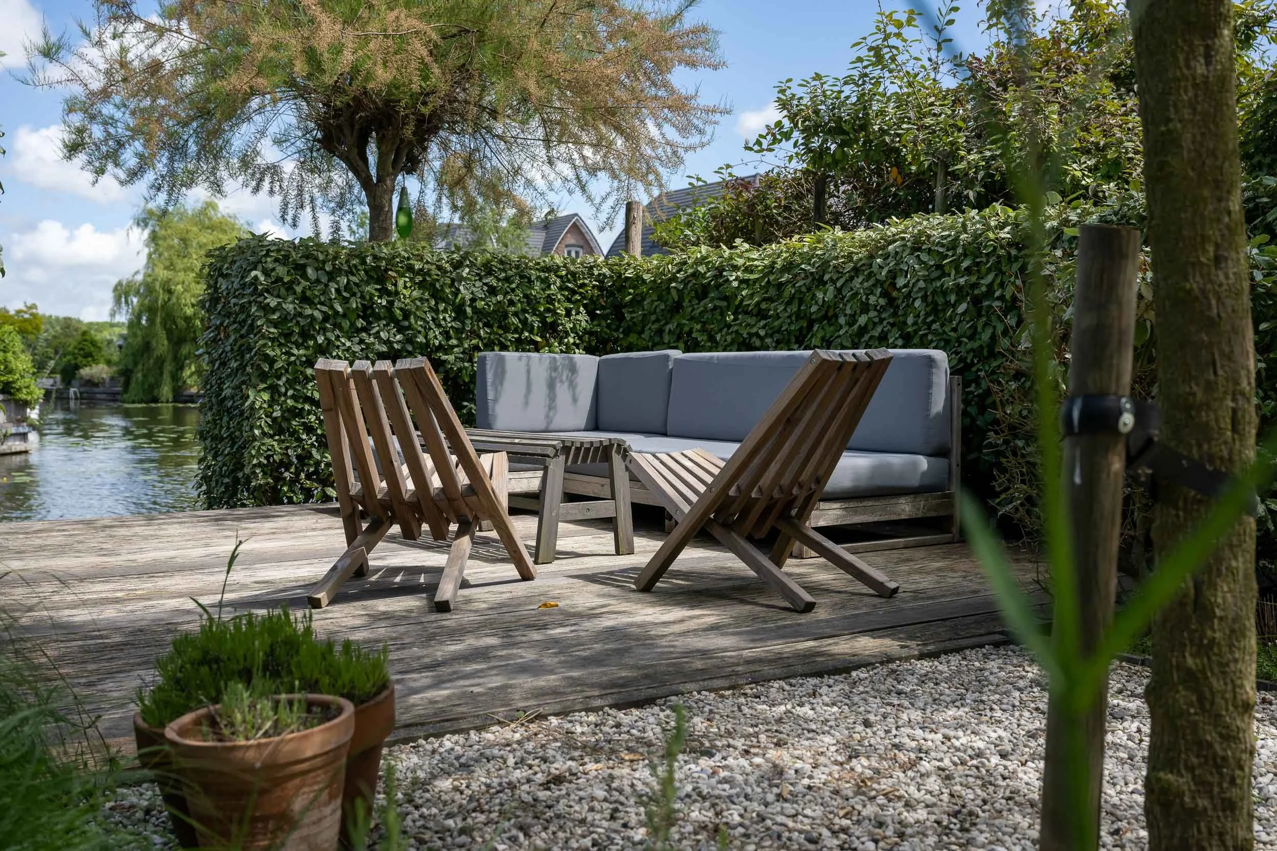 Een rustgevende tuin aan het water met houten meubelen, omgeven door hoge bomen en groen struikgewas.