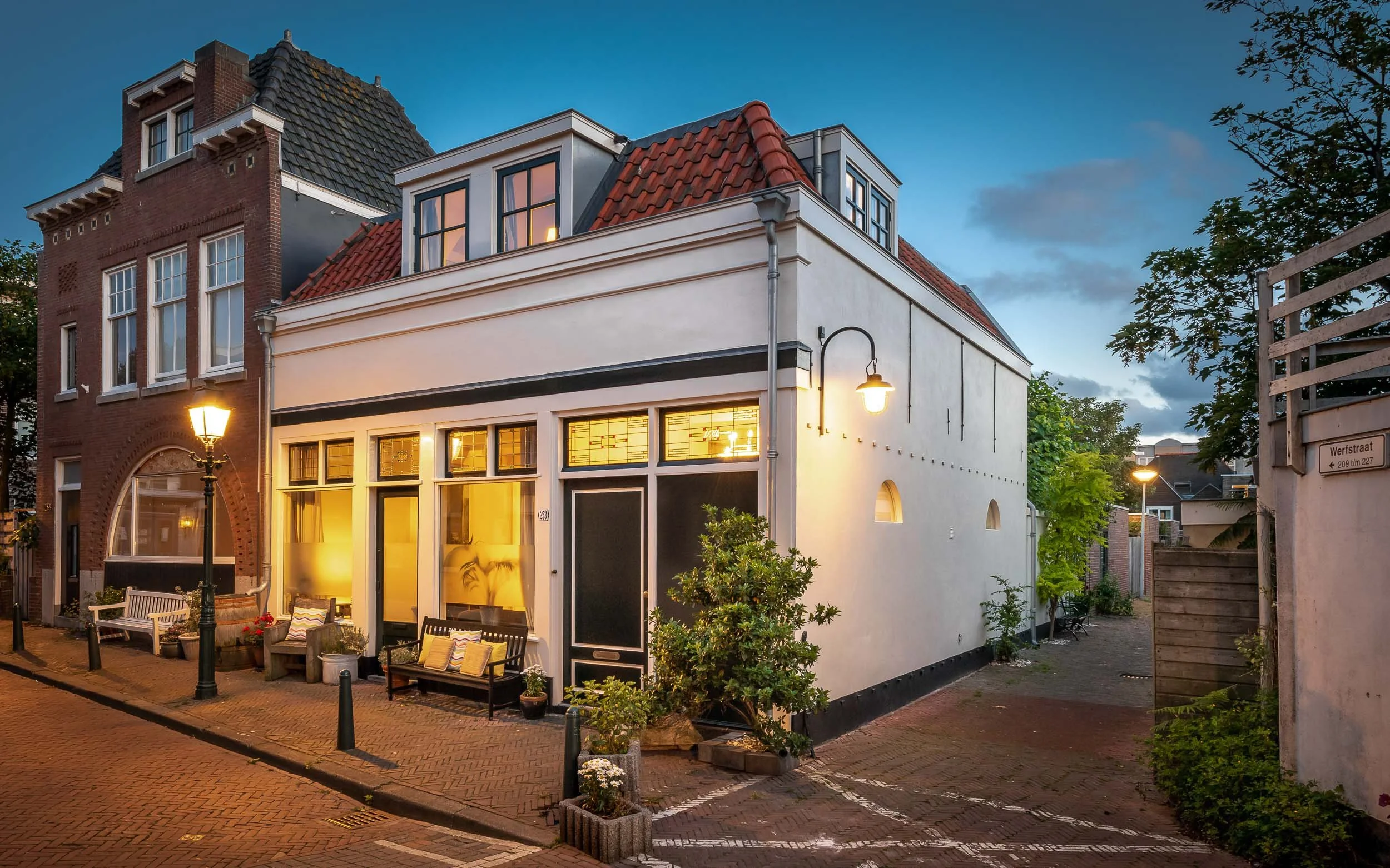 Gezellig huis met tuin bij schemering, buitenverlichting brandt, zitplaatsen en planten op straat.