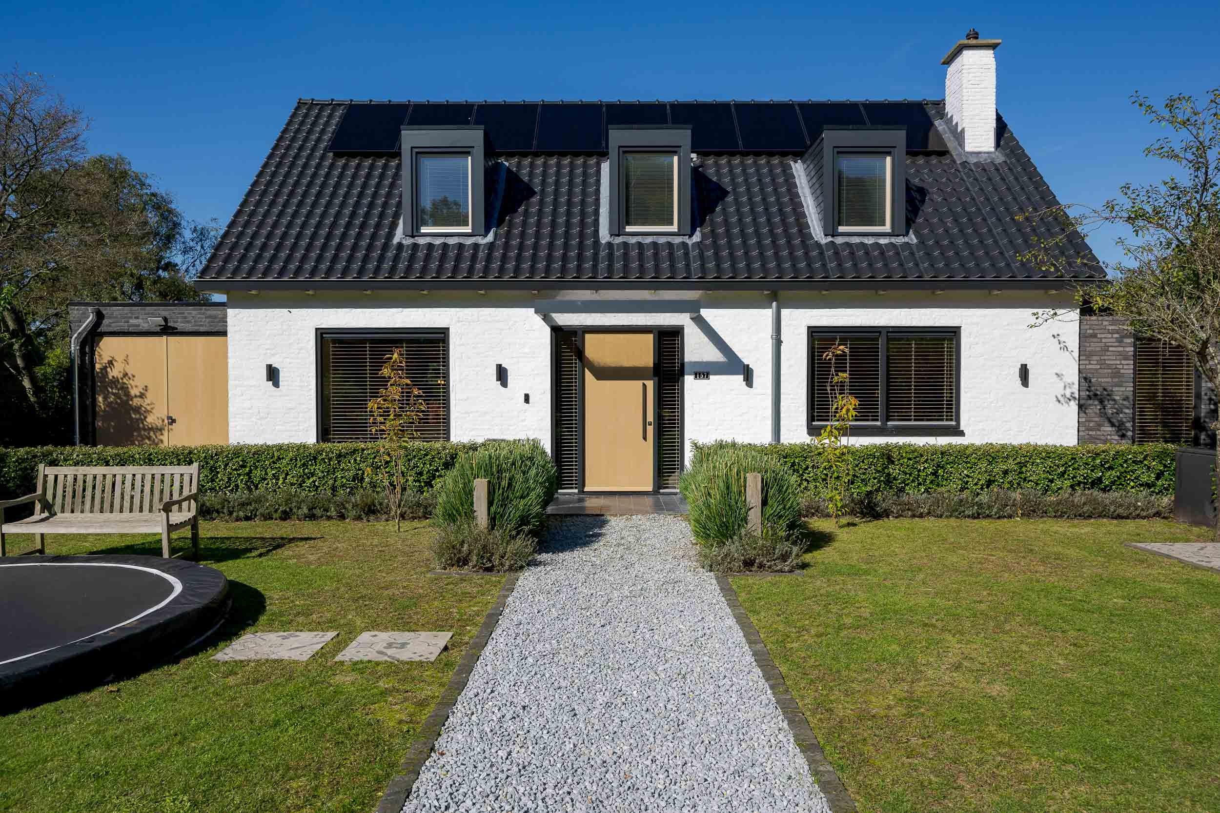Een moderne witte woning met zwarte dakpannen en drie dakkapellen. Voor de ingang ligt een grindpad dat naar de deur leidt, omringd door een groene tuin met struiken en een bank.