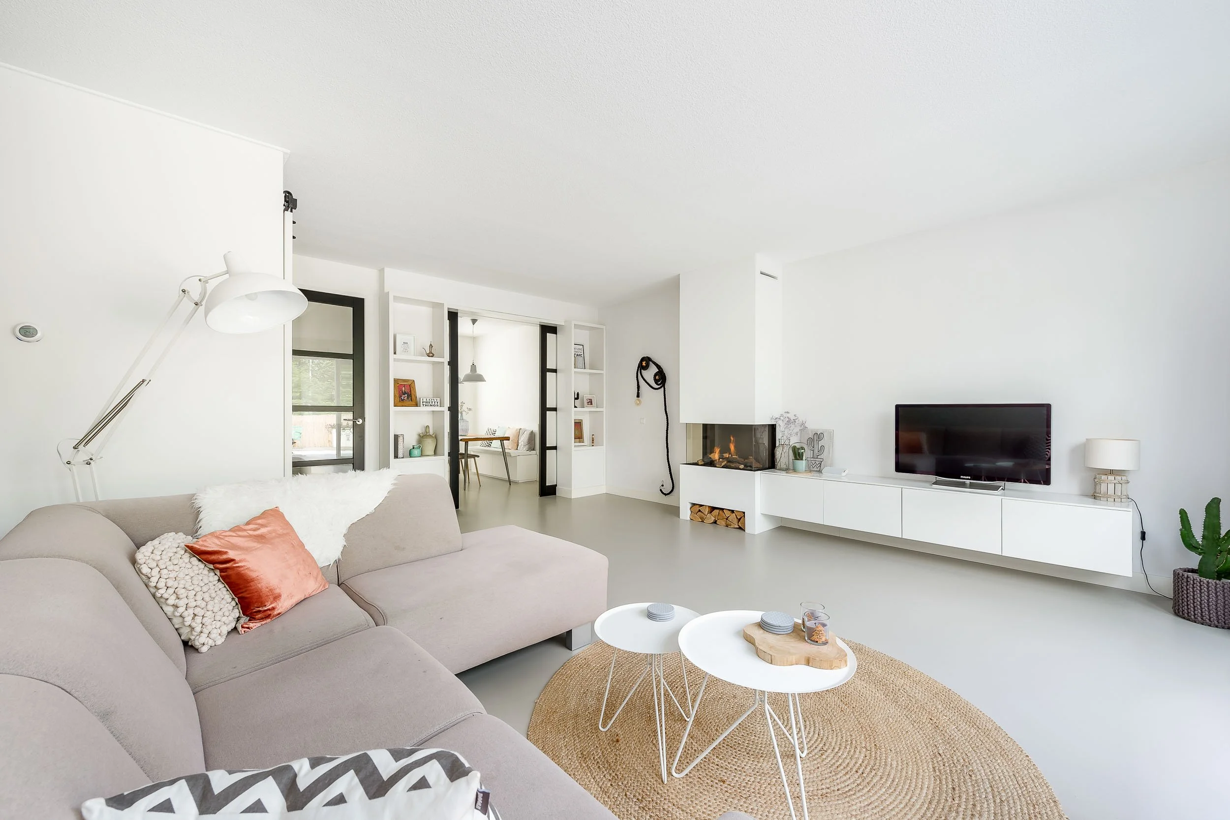 Hedendaagse woonkamer met een beige bank, witte salontafels op een mattenkleed, een open haard, een tv, en decoratie op planken en muren.