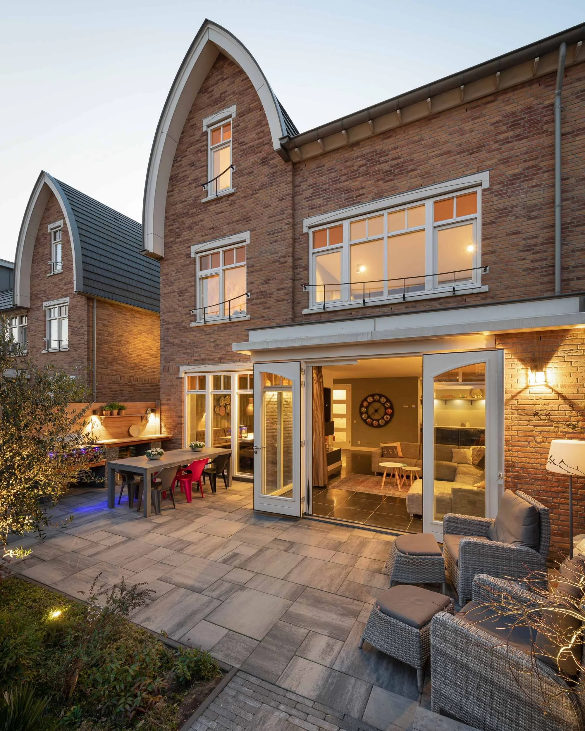 Uitzicht van een gezellige buitenruimte van een huis tijdens de avondschemering, met een terras met meubelen, een tuin met planten en een woning met grote ramen en veel binnenverlichting.