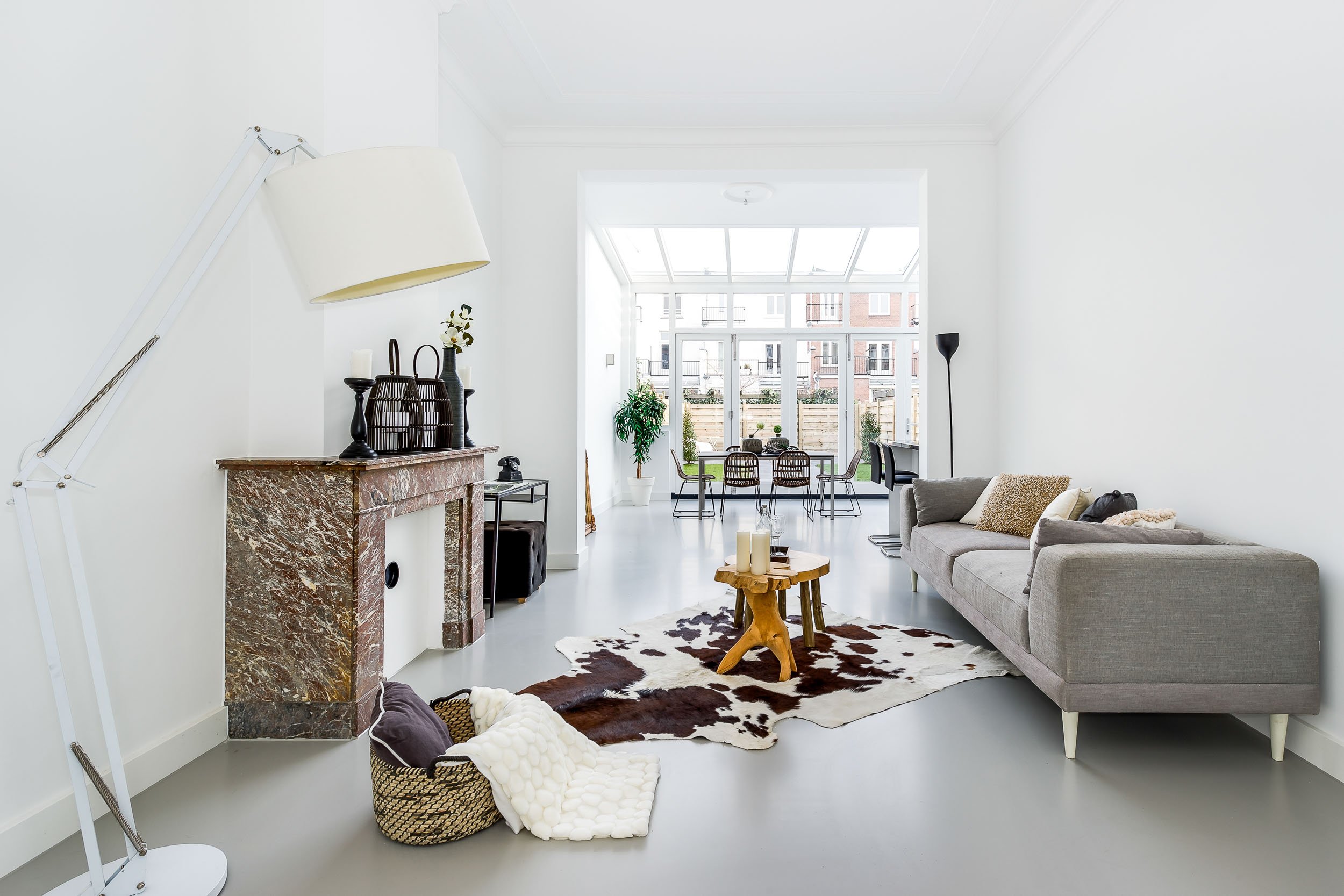 Een woonkamer met een grote raampartij, een loungebank, een tapijt met koeienprint, een kleine houten salontafel met kaarsen, een stenen schoorsteenmantel met decoratieve vazen en kandelaars, een vloerlamp en een tafel met eetstoelen.
