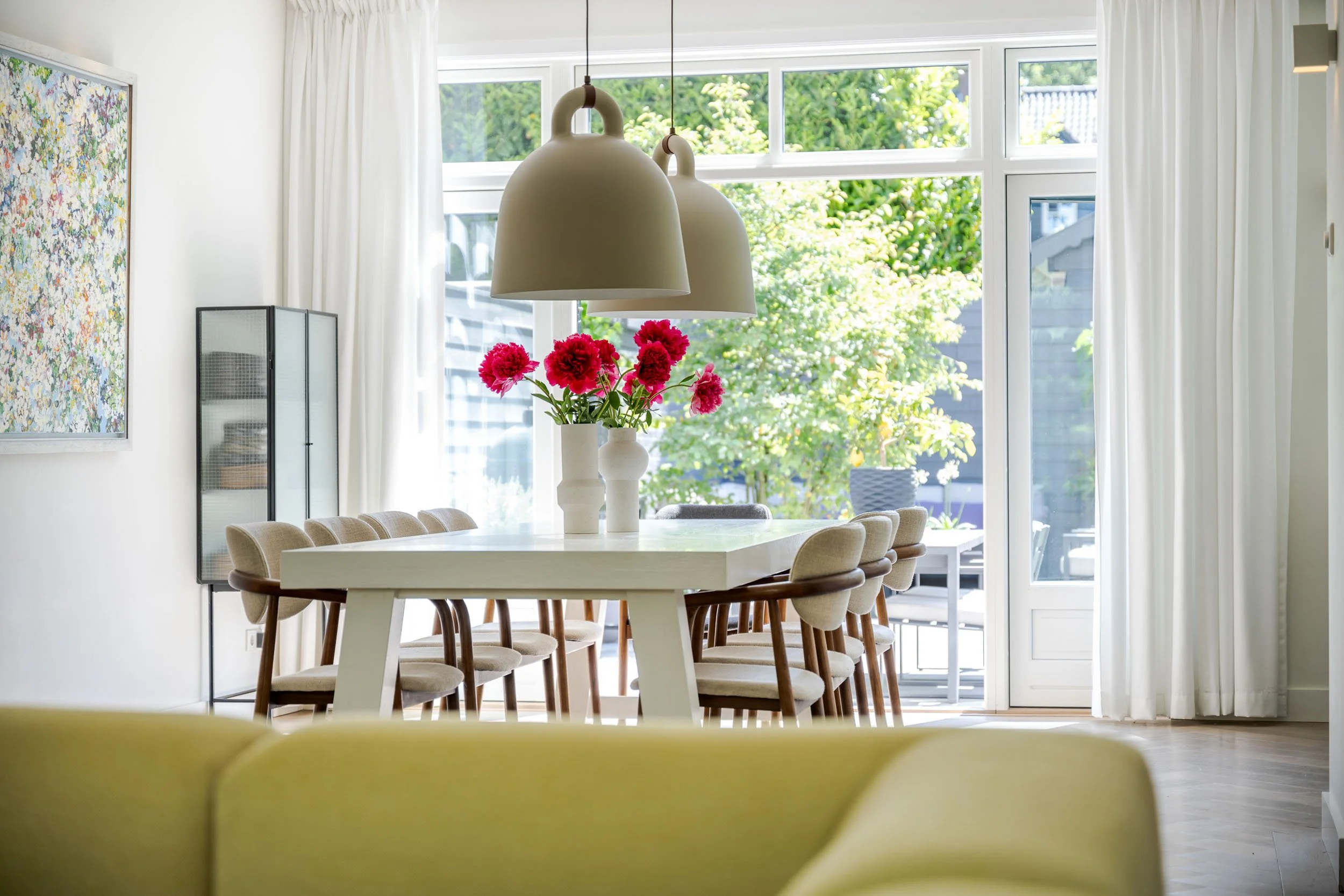 Eetkamertafel met twee vazen met roze bloemen, grote ramen met witte gordijnen, hanglampen boven de tafel, kleurrijk kunstwerk aan de muur, groene tuin buiten.