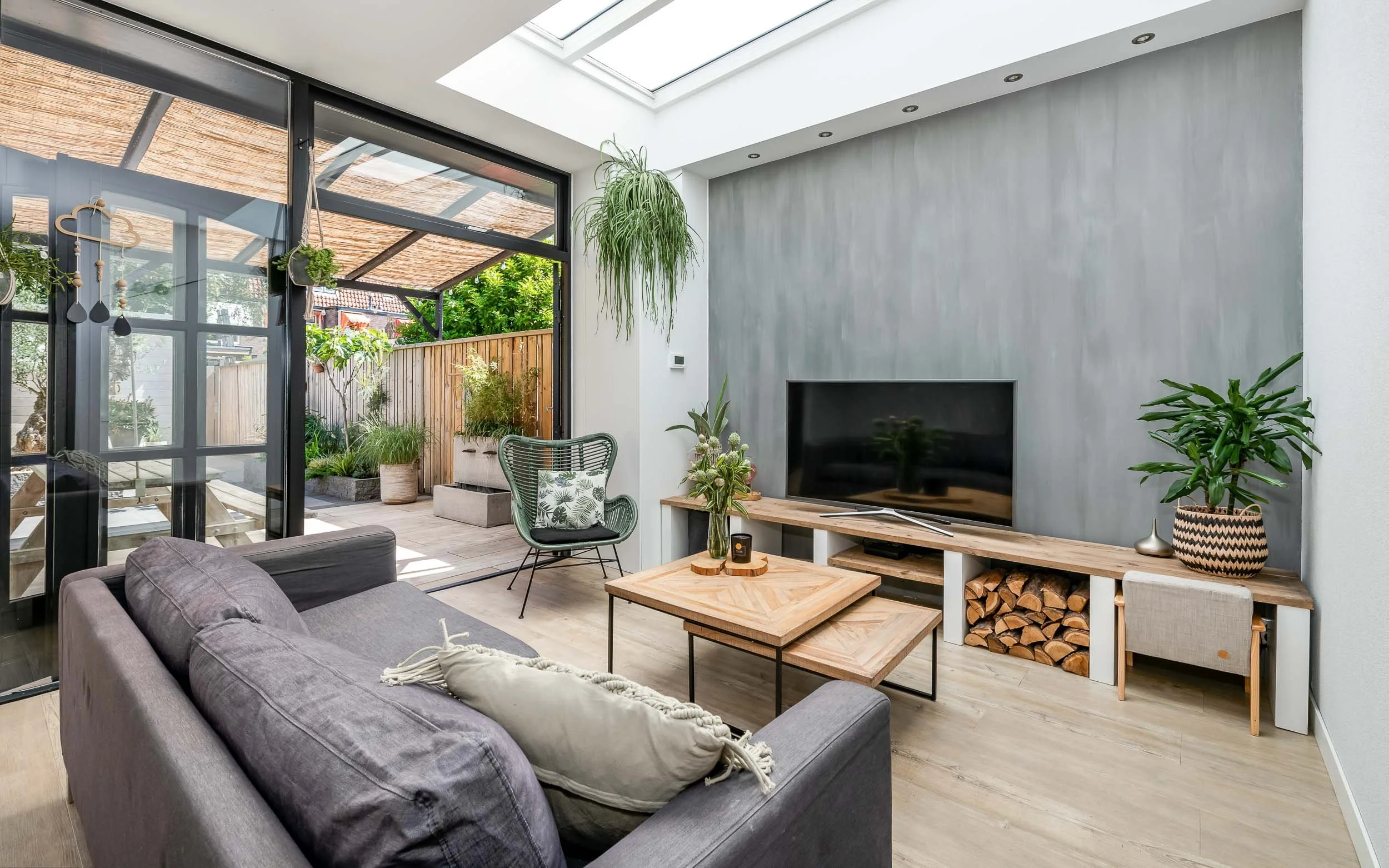 Woonkamer met grote glasdeuren die leiden naar een tuin, met een tv, planten, en een houten salontafel.