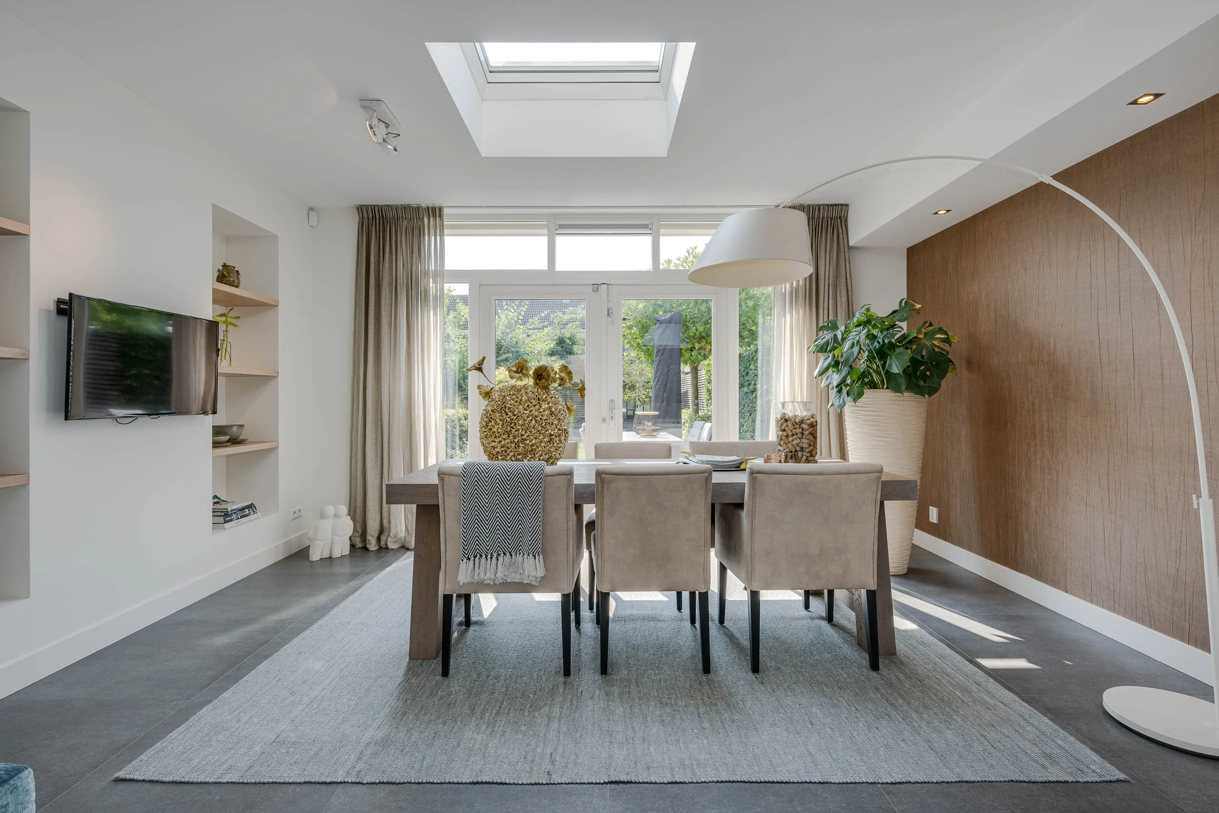 Interieur van een woonkamer met een eettafel, zes stoelen, een grote plantenbak, een vloerlamp, een tv, een raam, een plafondlicht en een dakraam.