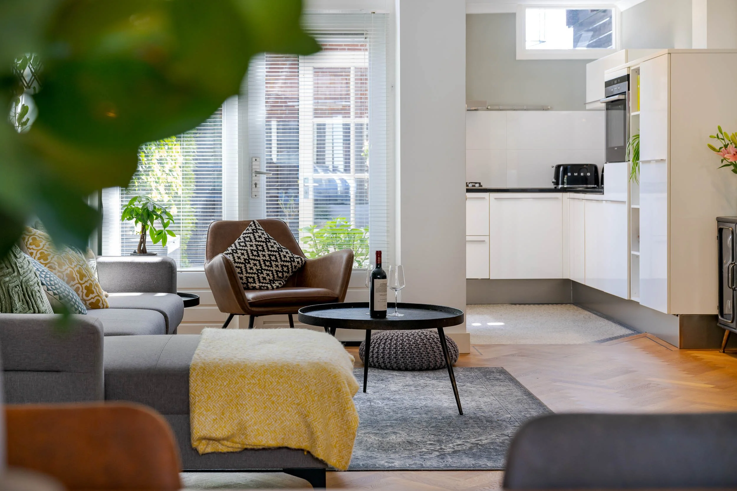 Woonkamer met zithoek, planten, grote ramen met blinds, en een open keuken met wit keukenmeubilair en apparaten.