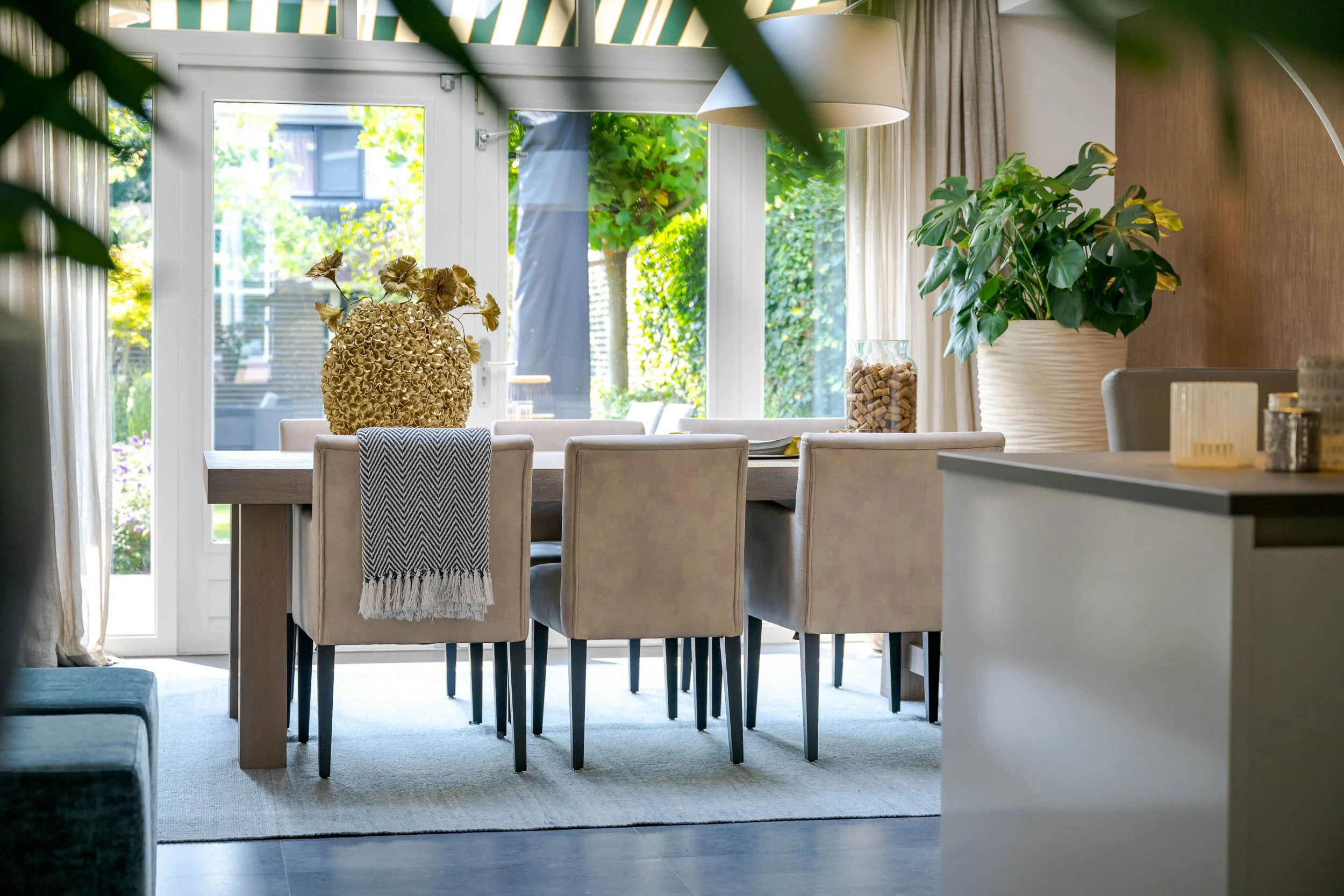 Binnenruimte met een grote tafel, omringd door beige stoelen, met een grote vaas met gedroogde bloemen, een grote groene plant in een witte pot en een glazen pot met kurken, met grote ramen en gordijnen.