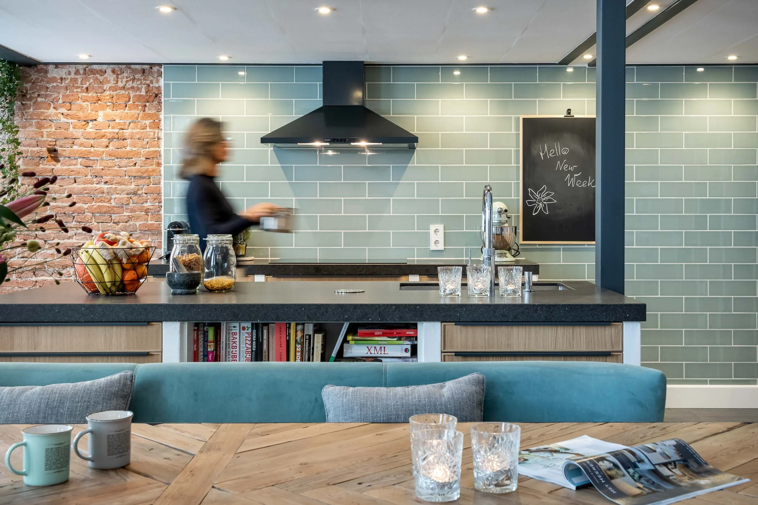 Een moderne keuken van een woning in Voorschoten met een bakstenen muur en mintgroene tegels, een zwart afzuigkap en een eettafel met boeken, kopjes en gelezen tijdschriften op de voorgrond.