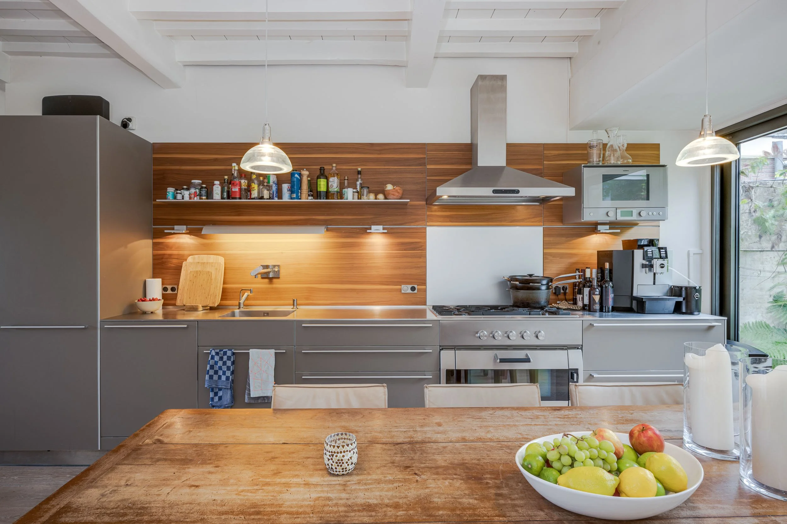 Moderne keuken met houten en grijze afwerkingen, open schappen met kruiden en etenswaren, ingebouwde oven, en een tafel met een kom fruit.