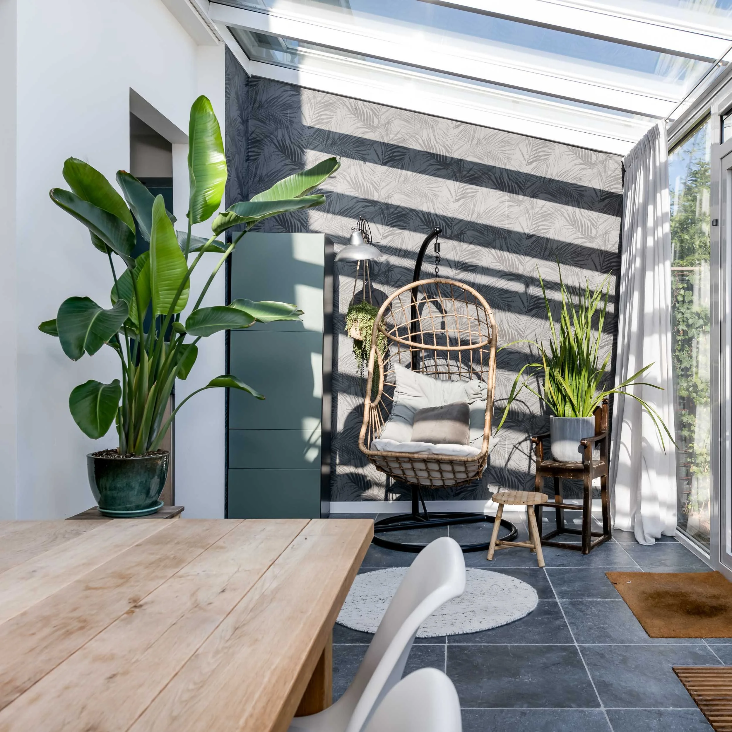 Binnenruimte met grote plantenpot met groene bladeren, een houten tafel, witte stoel, een hangmat en een zithoek bij een raam met vitrage, en een muur met een botanisch patroon op de achtergrond.