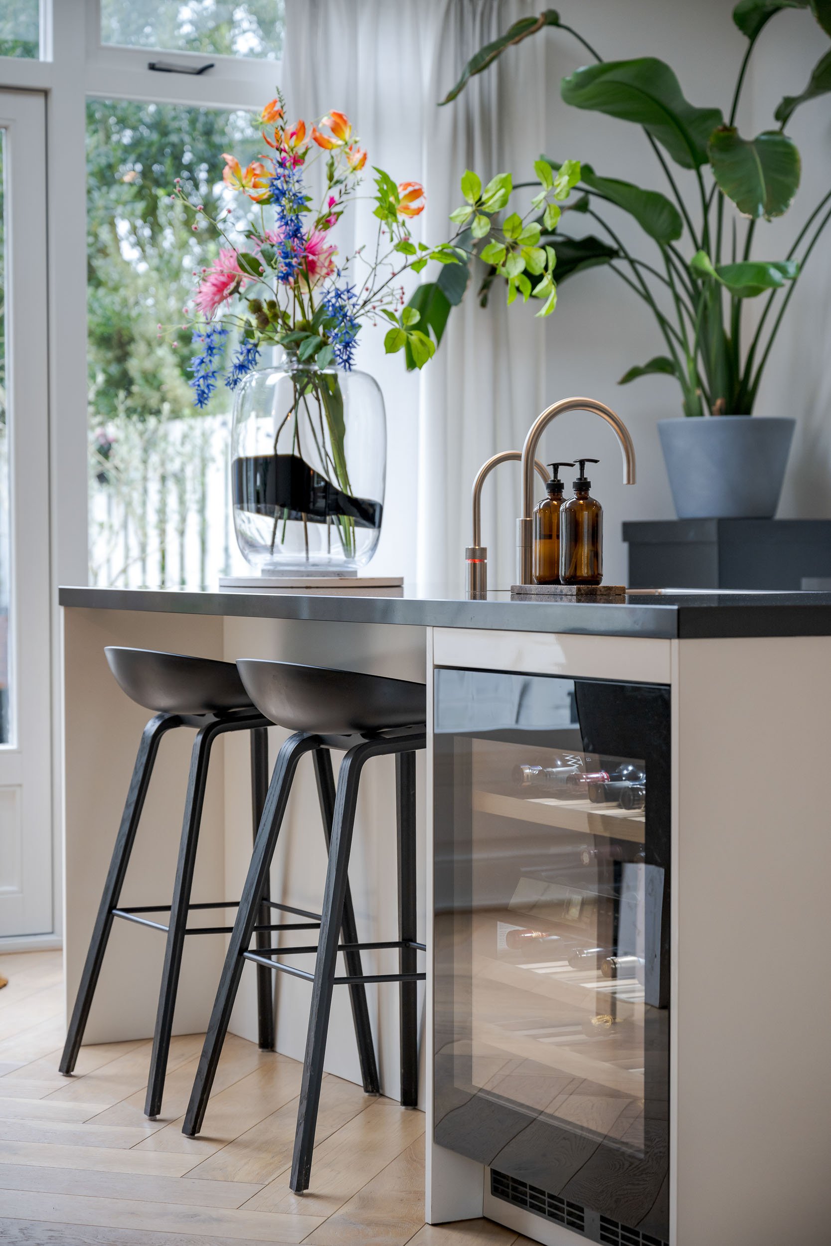 Een keuken met een ontbijtbar met drie zwarte barkrukken en planten. Op de achtergrond een keuken met een gootsteen en een wijnkoelkast onder het aanrecht. Er staat een vaas met een kleurrijk boeket bloemen.