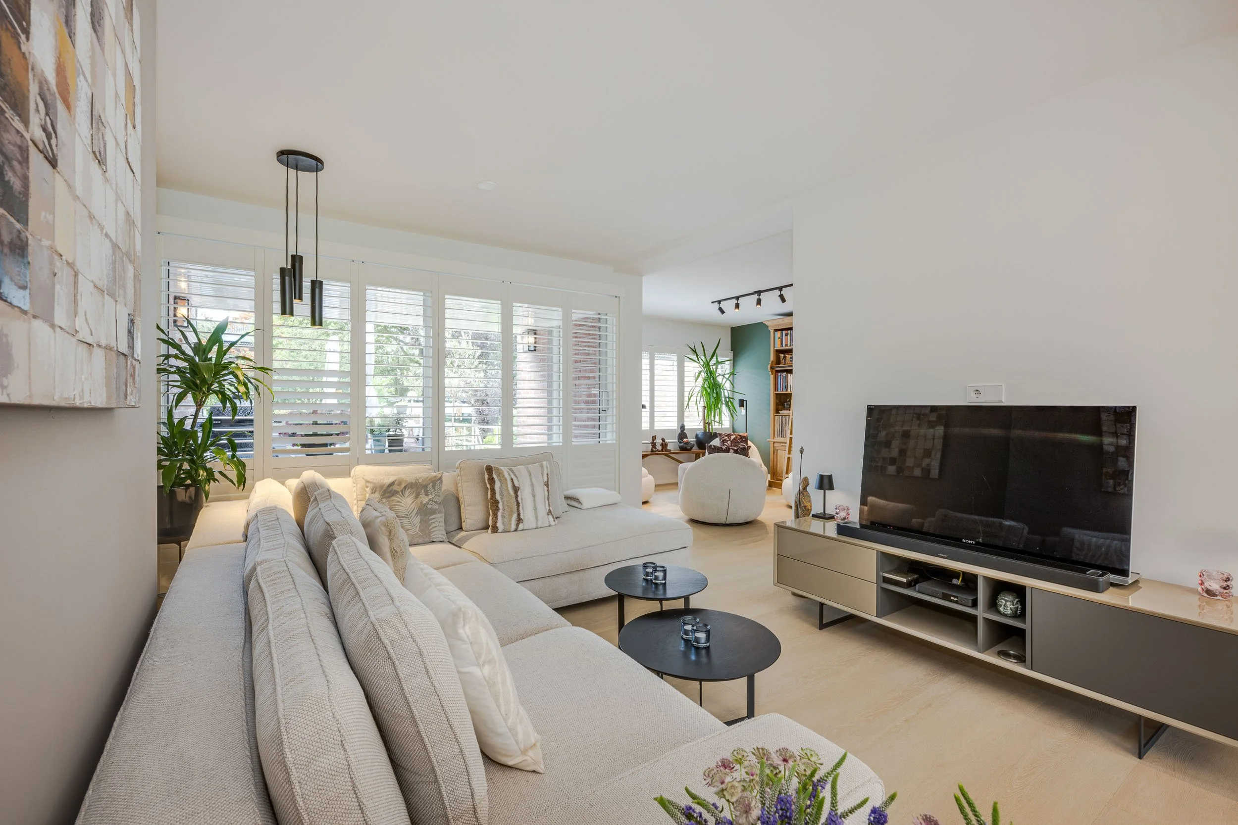 Woonkamer met witte bank, ronde zwarte salontafels, grote tv, planten, boekenplank, grote ramen met shutters, veel natuurlijk licht.
