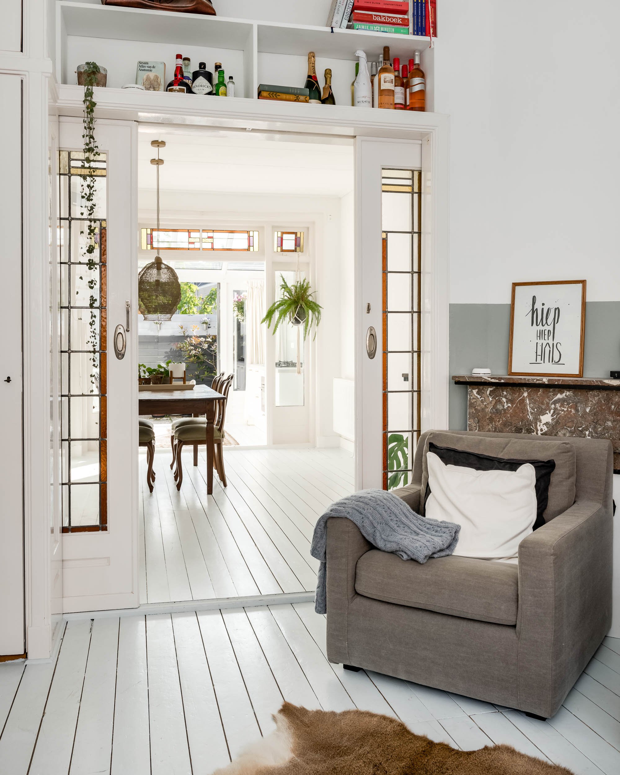 Woonkamer met grijze fauteuil, witte muur, schilderij met tekst 'hiep hiep hoera', groene planten, eettafel met stoelen, en decoratieve ramen met gekleurde glas-in-lood details. Glazen deur leidt naar een lichte tuin.