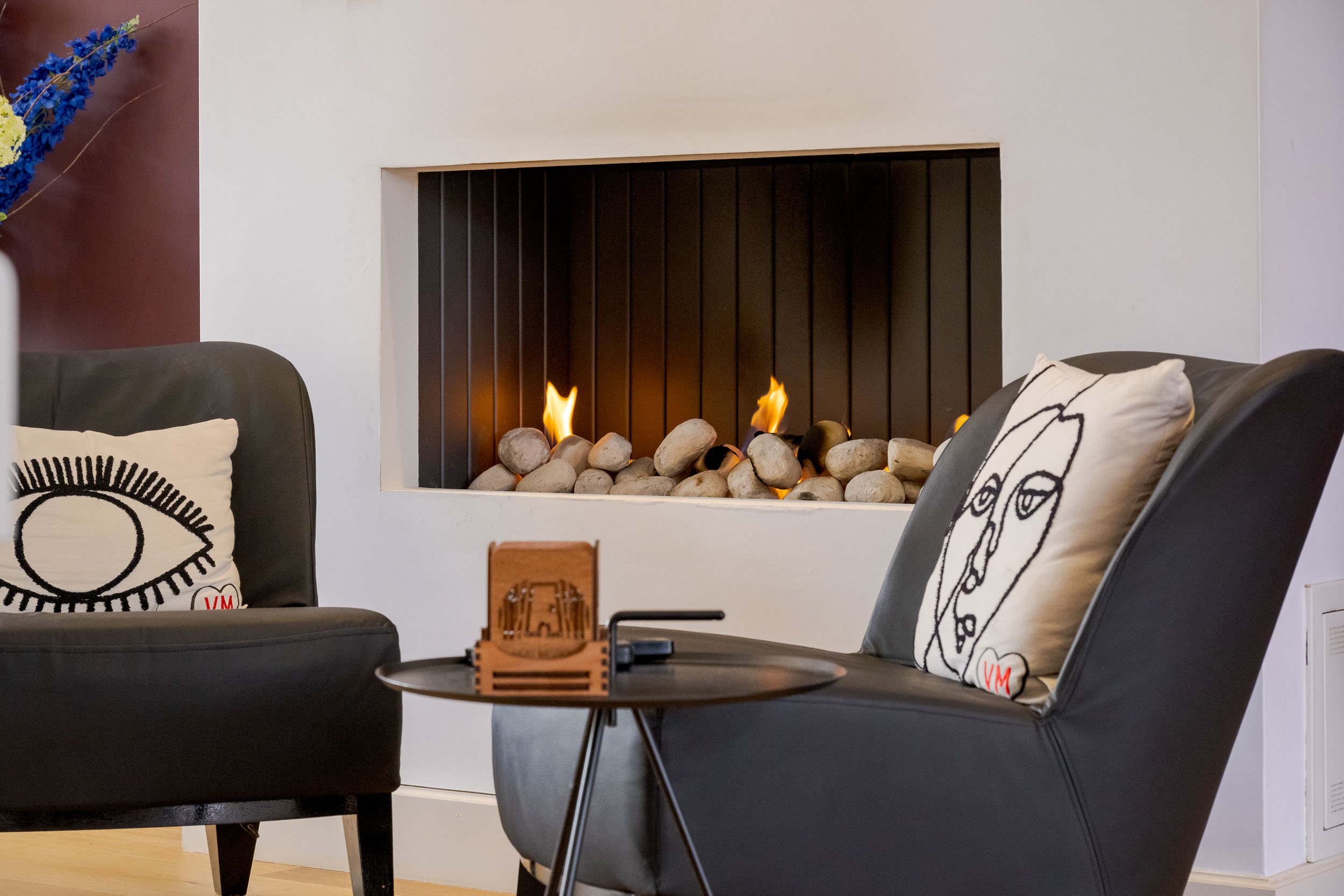 Woonkamer met moderne elektrische haard, zittende stoelen met kussens en bloemen in vaas.