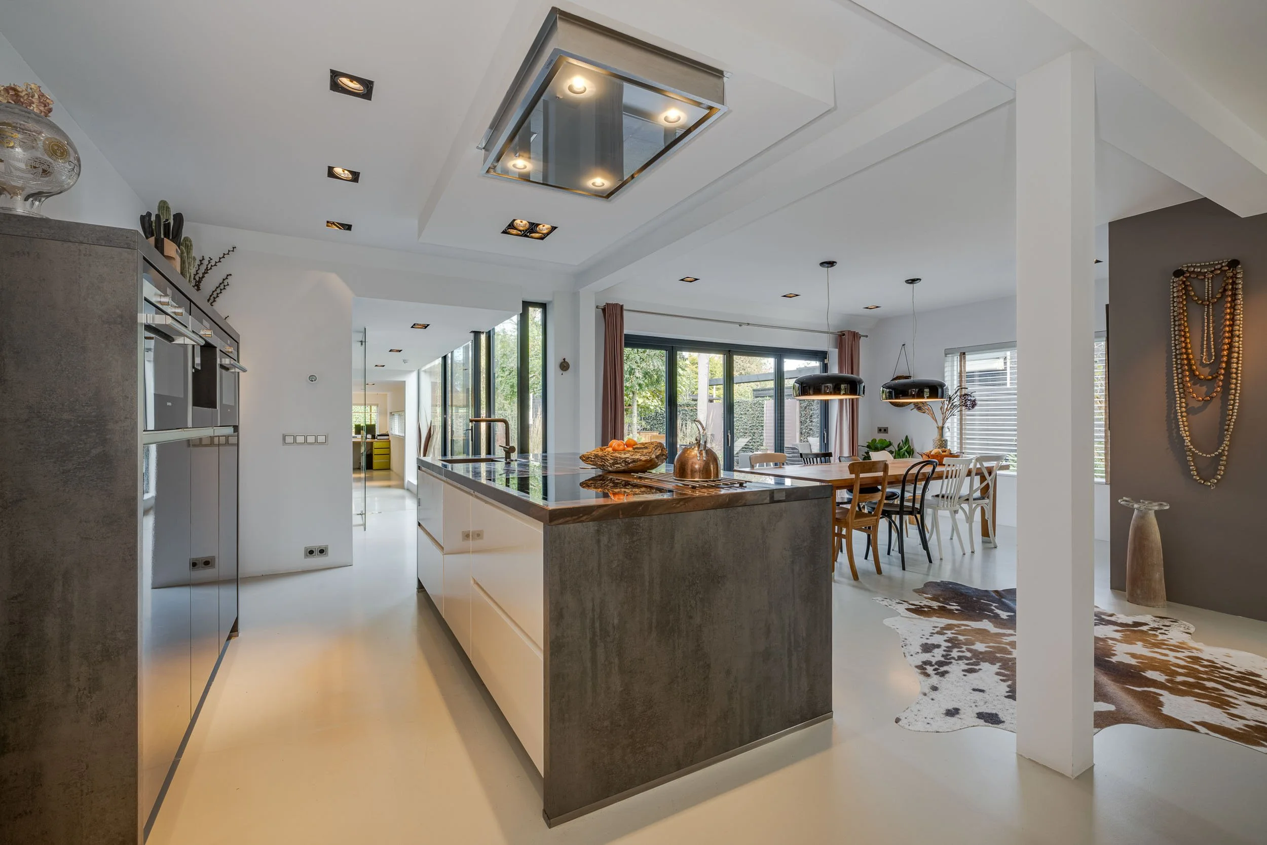 Moderne open keuken met een centraal ontbijt- of keukeneiland, grote ramen die natuurlijk licht binnenlaten, en een zithoek met eetkamertafel en stoelen.