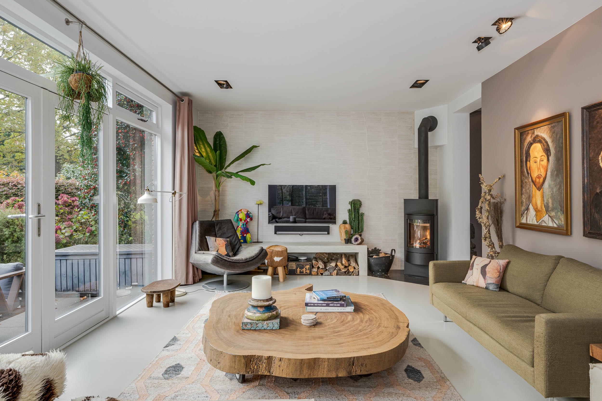 Woonkamer met grote ramen, planten, houtkachel, bank en kunstwerken.