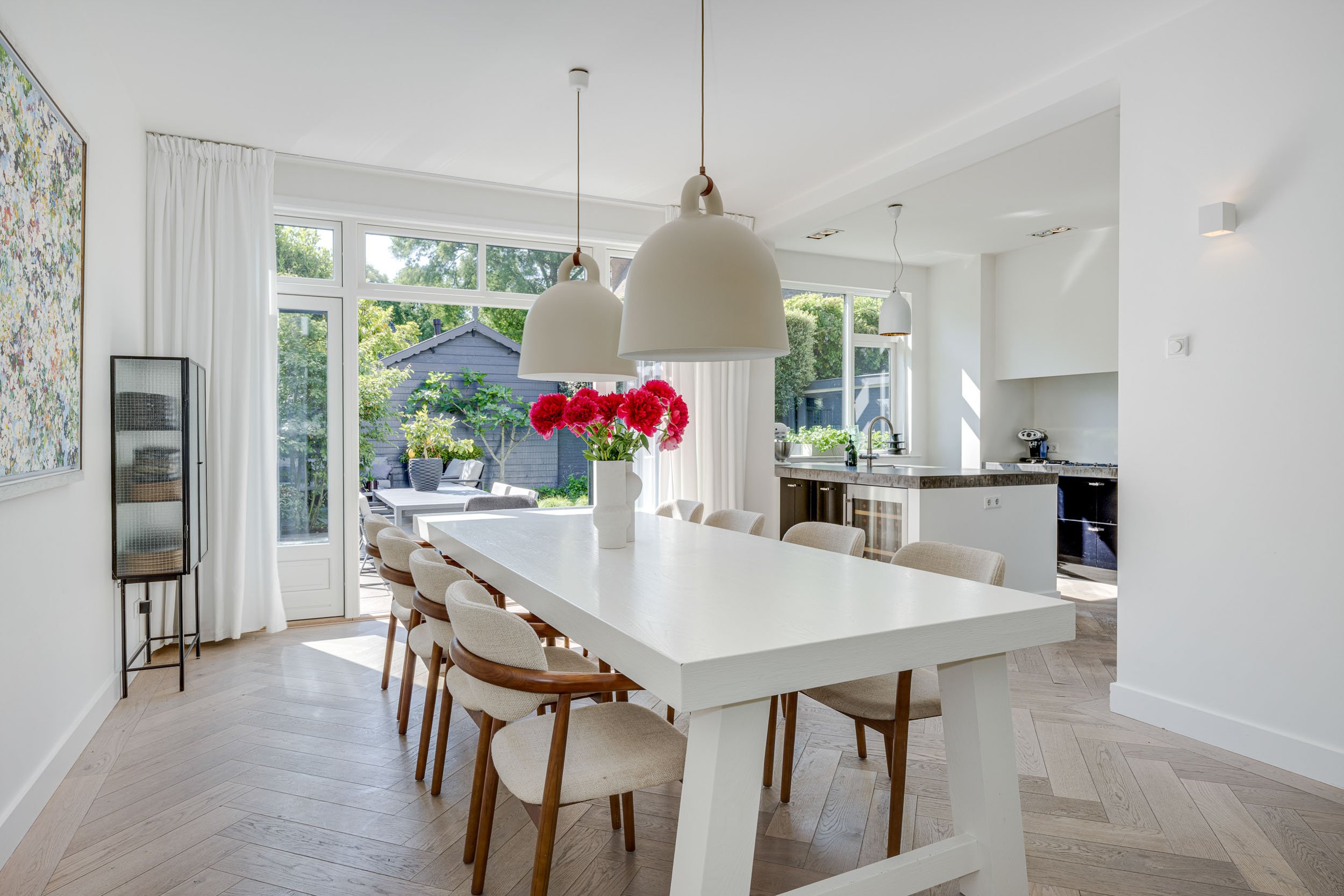Witte eetkamer met lange tafel, beige stoelen, bloemen in vaas, grote ramen en uitzicht op tuin in een grote villa in Wassenaar.