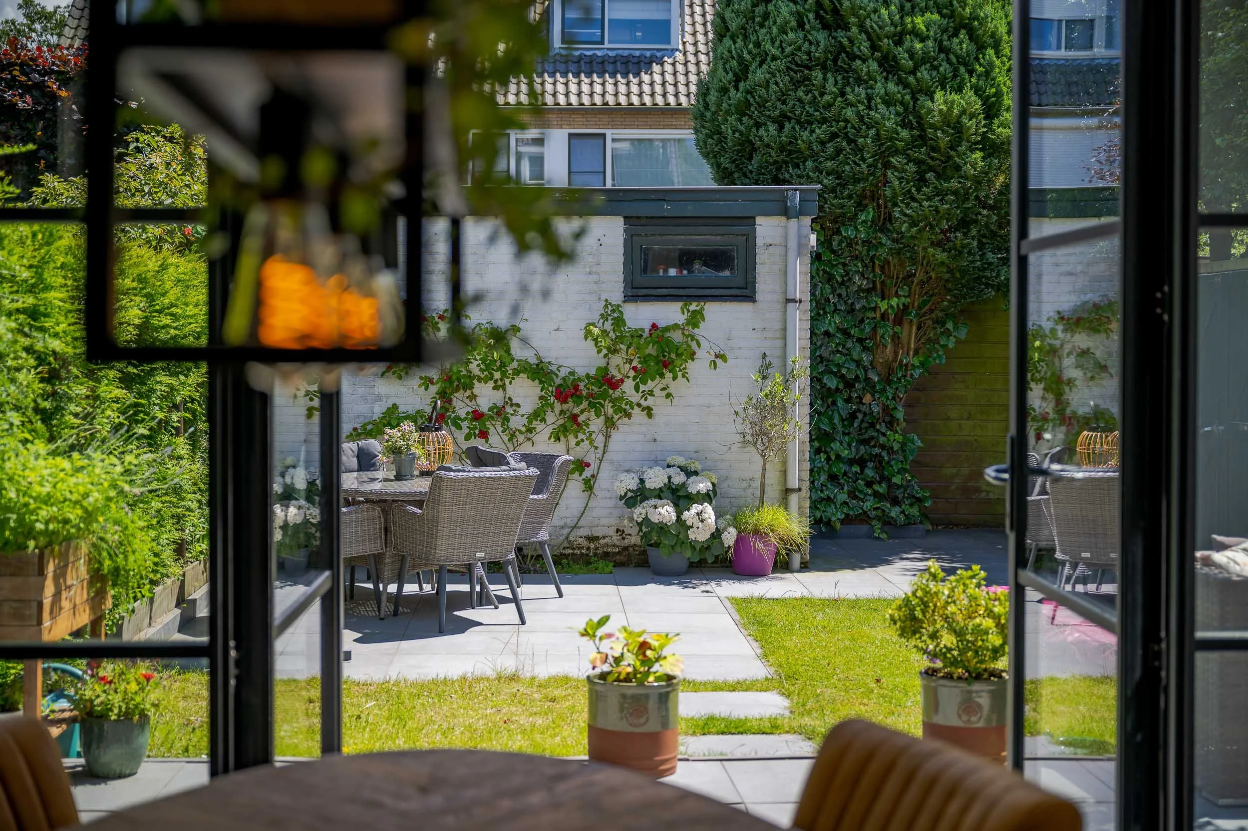 Uitzicht op een tuin gezien vanuit binnen, met een eettafel, tuinstoelen en verschillende planten, bomen en een gebouw op de achtergrond.