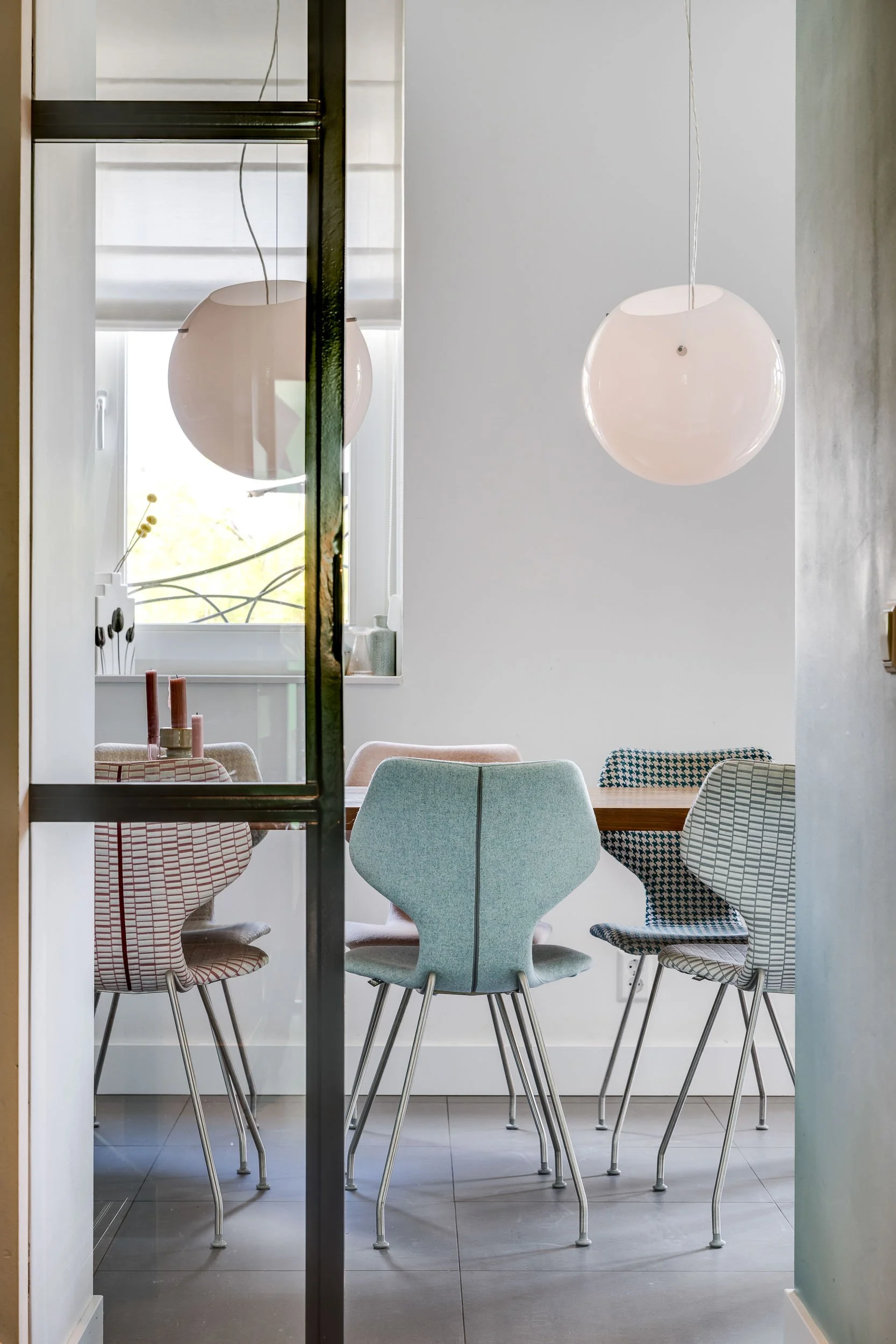 Een eetkamer met zes kleurrijke stoelen rond een houten tafel, een raam met witte jaloezieën, en hanglampen boven de tafel.