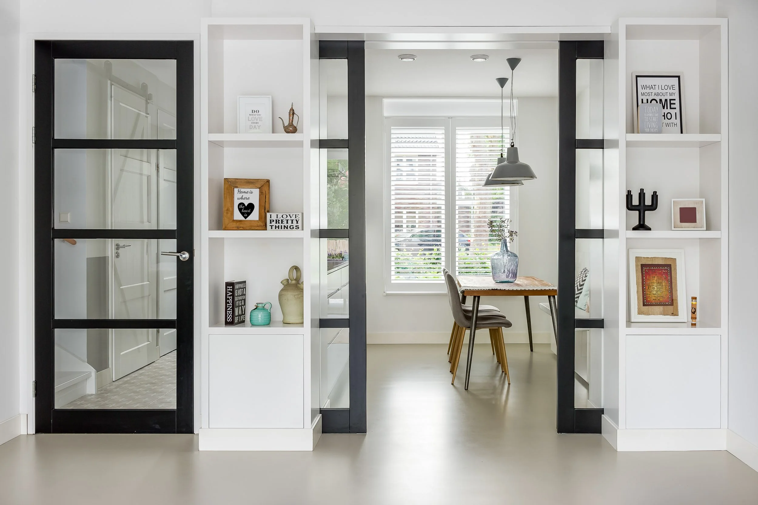 Interieur van een modern huis met witte en zwarte muurrekken, een tafel met een vaas met bloemen, en grote ramen met luiken.