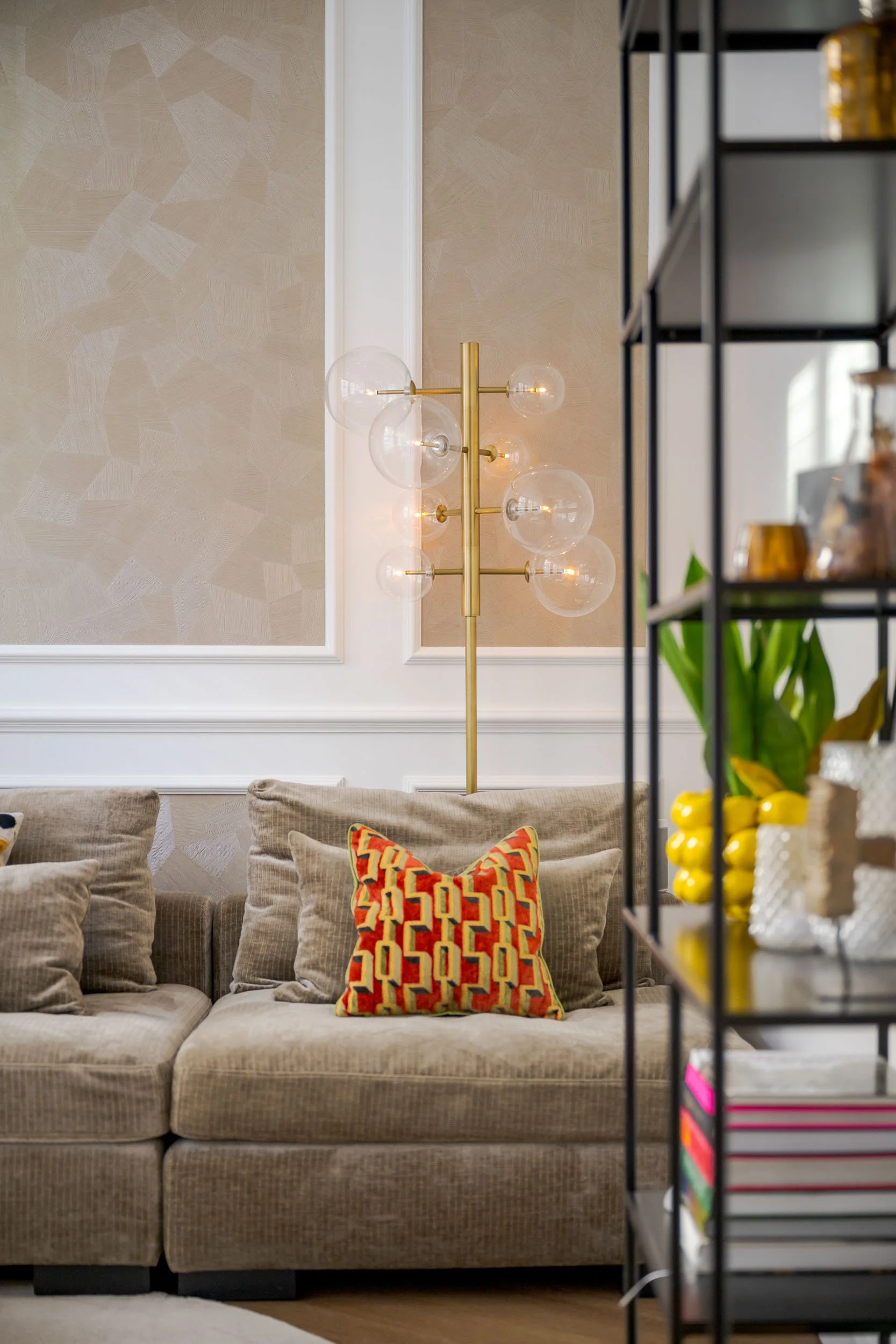 Gezellig huisinterieur met een beige bank, kleurrijk kussen, modern plafondlamp en open glazen boekenkast gevuld met boeken en decoraties.