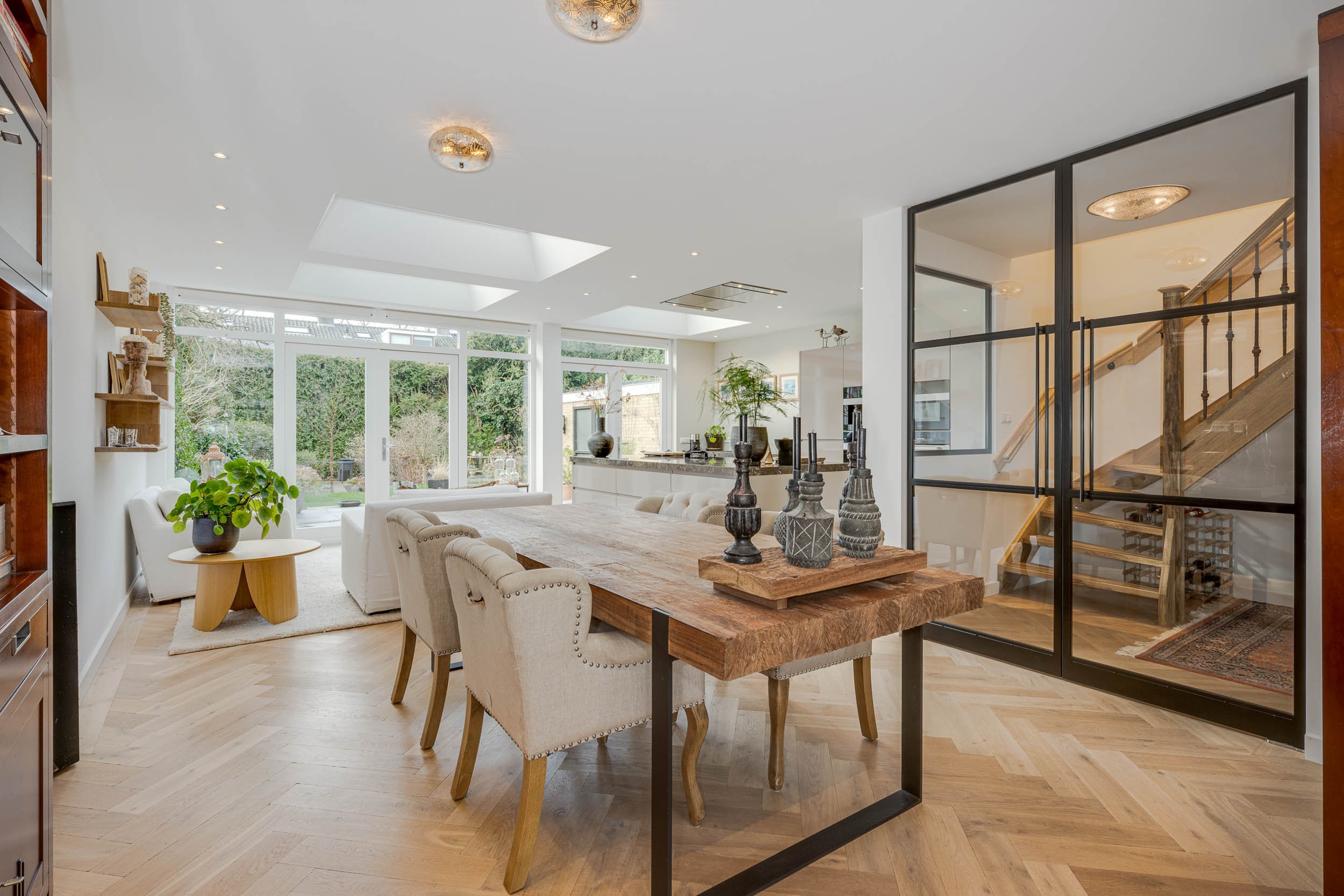 Ruimtelijk interieur van een woonkamer met grote ramen, een eettafel met decoratieve vazen, een zithoek met witte banken, houten vloeren, en een stalentrap.