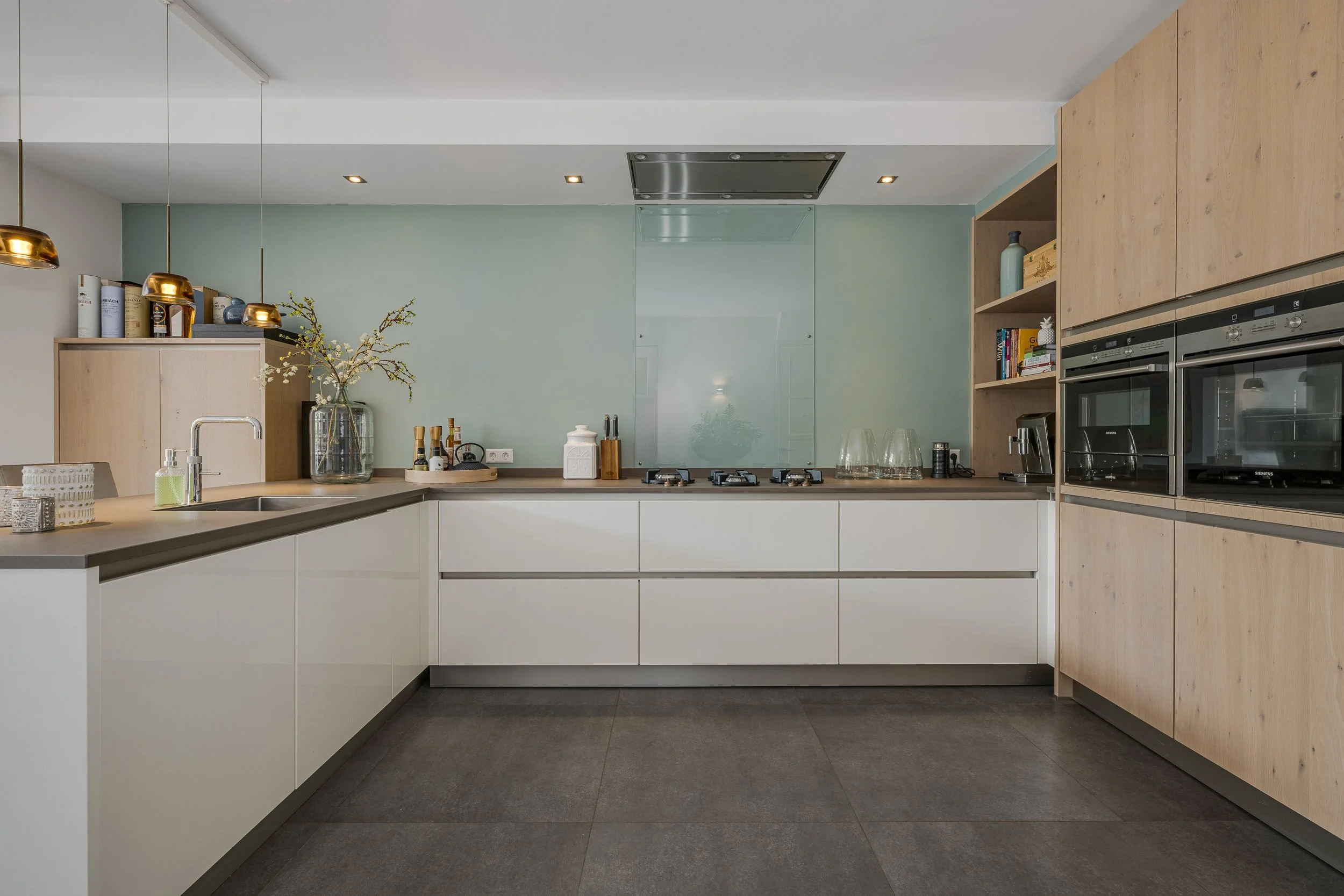 Moderne keuken met witte kasten, houten wandkast, ingebouwde oven en vaatwasser, gasfornuis, groene muur, en decoratieve objecten zoals vazen en boeken.