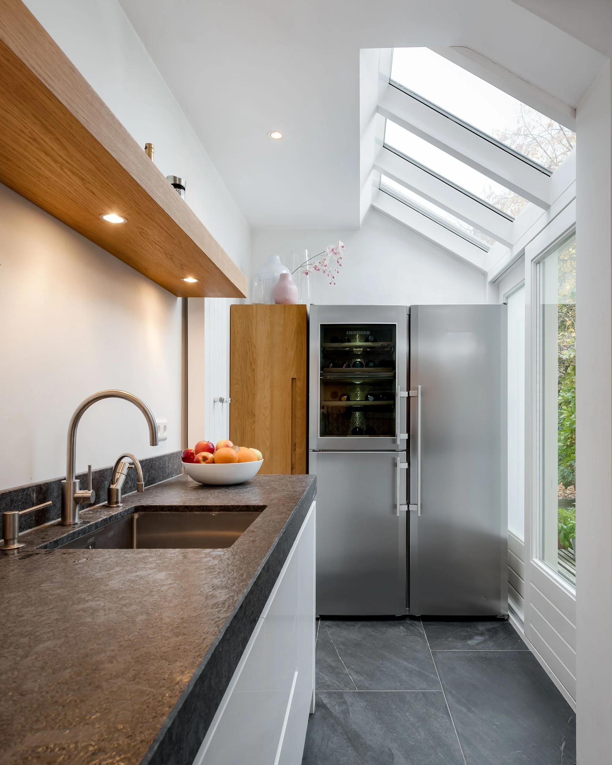 Moderne keuken met een granito werkblad, een waterkraan en een fruitschaal met appels, met een grote koelkast en een venster. Boven de keuken hangt een houten plank met decoratieve vazen en een lichtspot.